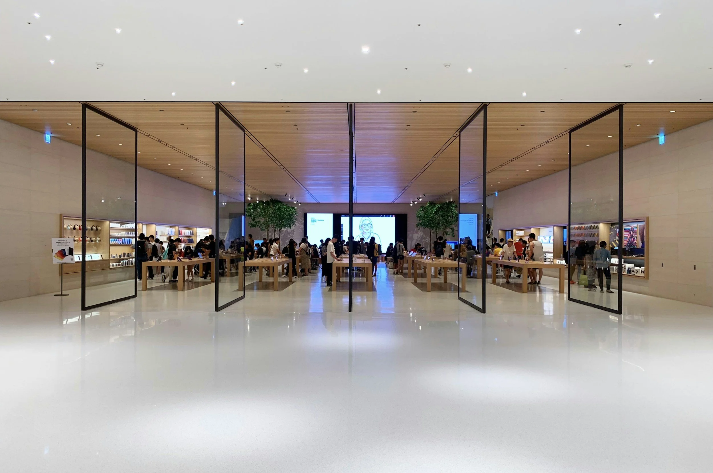 Inside view of a modern retail store with people shopping and interacting with electronic devices, with wooden ceiling, display shelves, and digital screens on the walls.