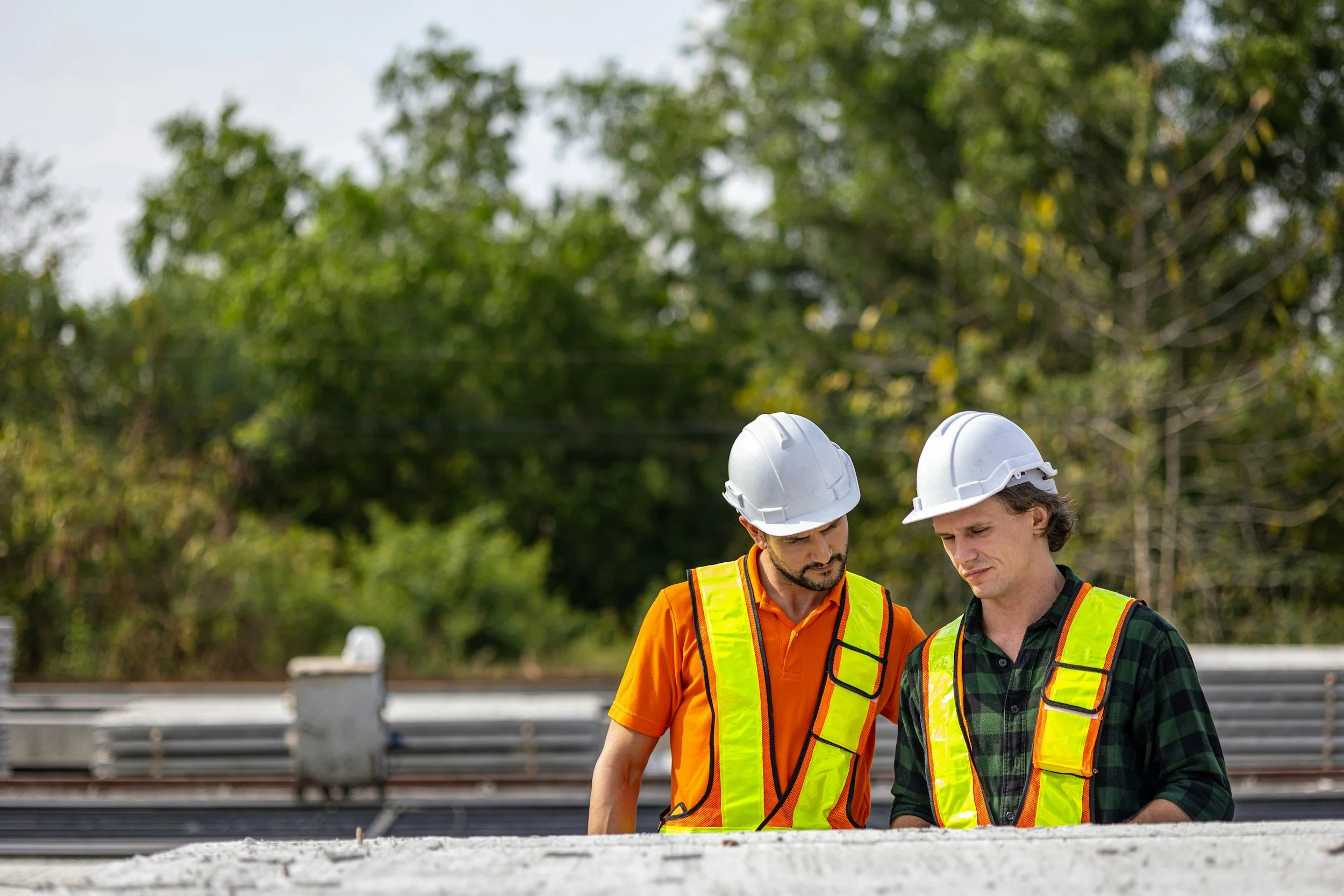 Two construction workers wearing hard hats and safety vests standing outdoors near a construction site with trees in the background.