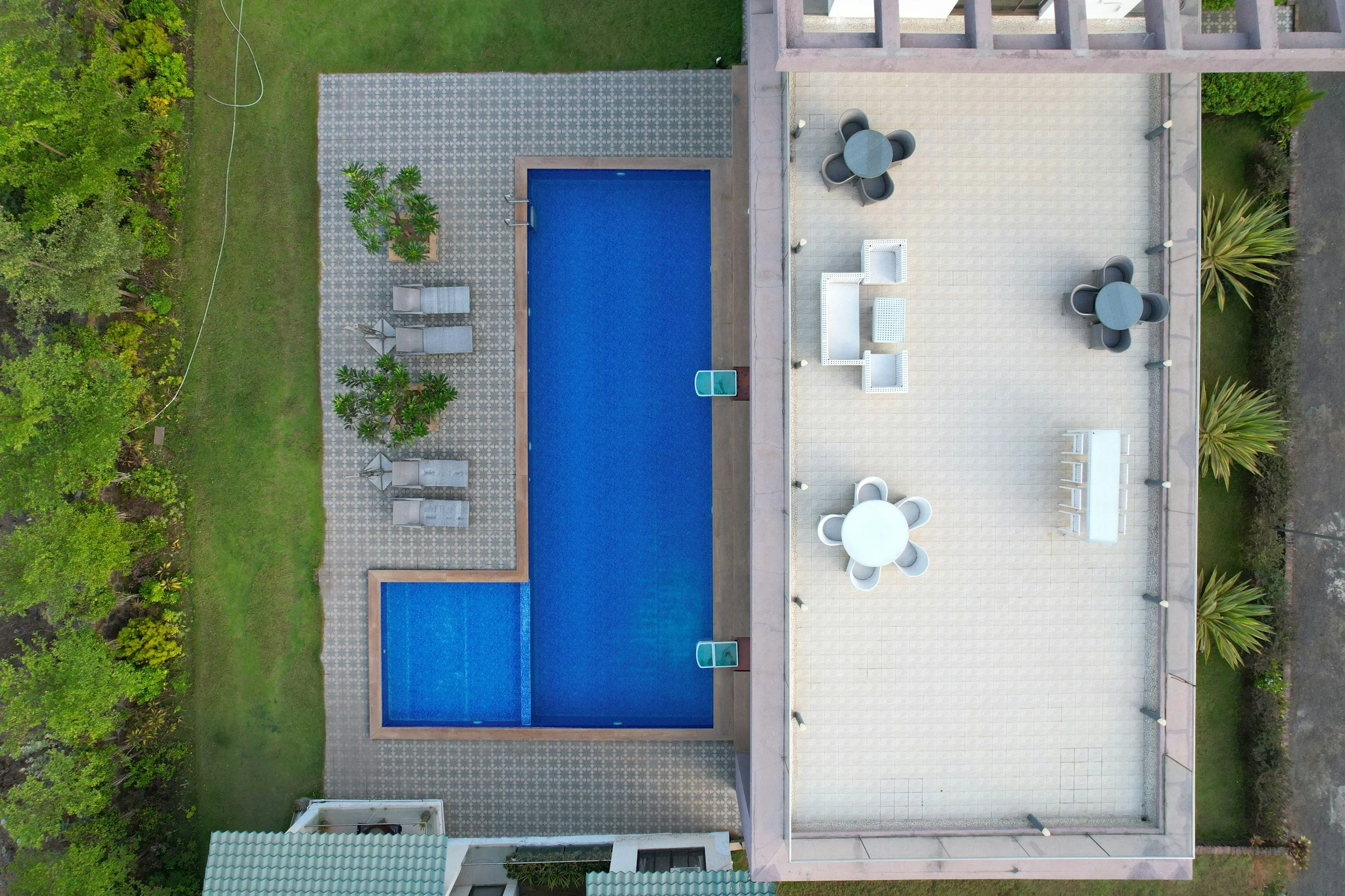 An aerial view of a backyard showing a swimming pool, surrounded by a tiled patio area with outdoor furniture, and lush green trees on one side.