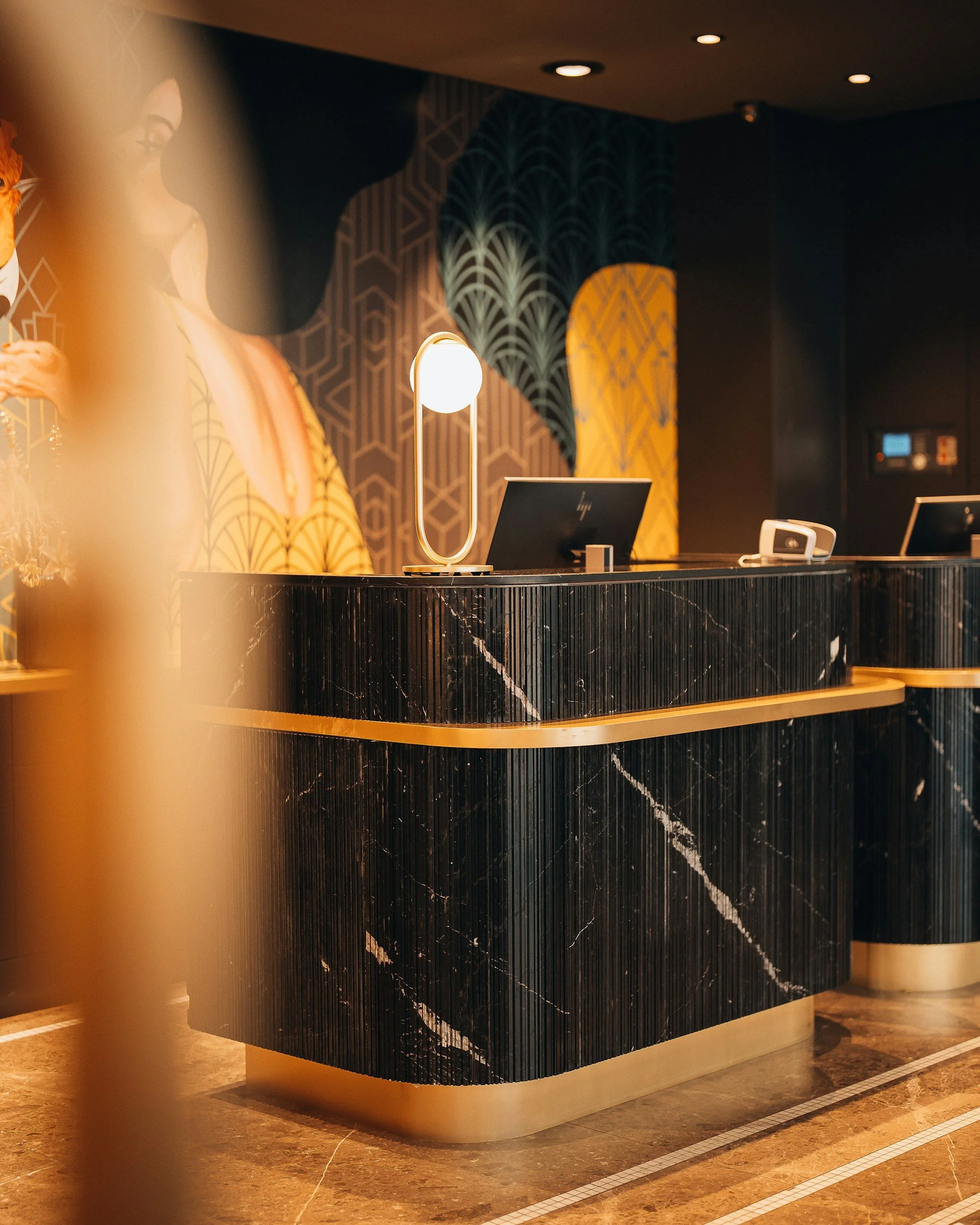 Luxurious hotel reception area with a curved black marble front desk, illuminated lamp on top, and patterned wallpaper in gold and dark colors in the background.