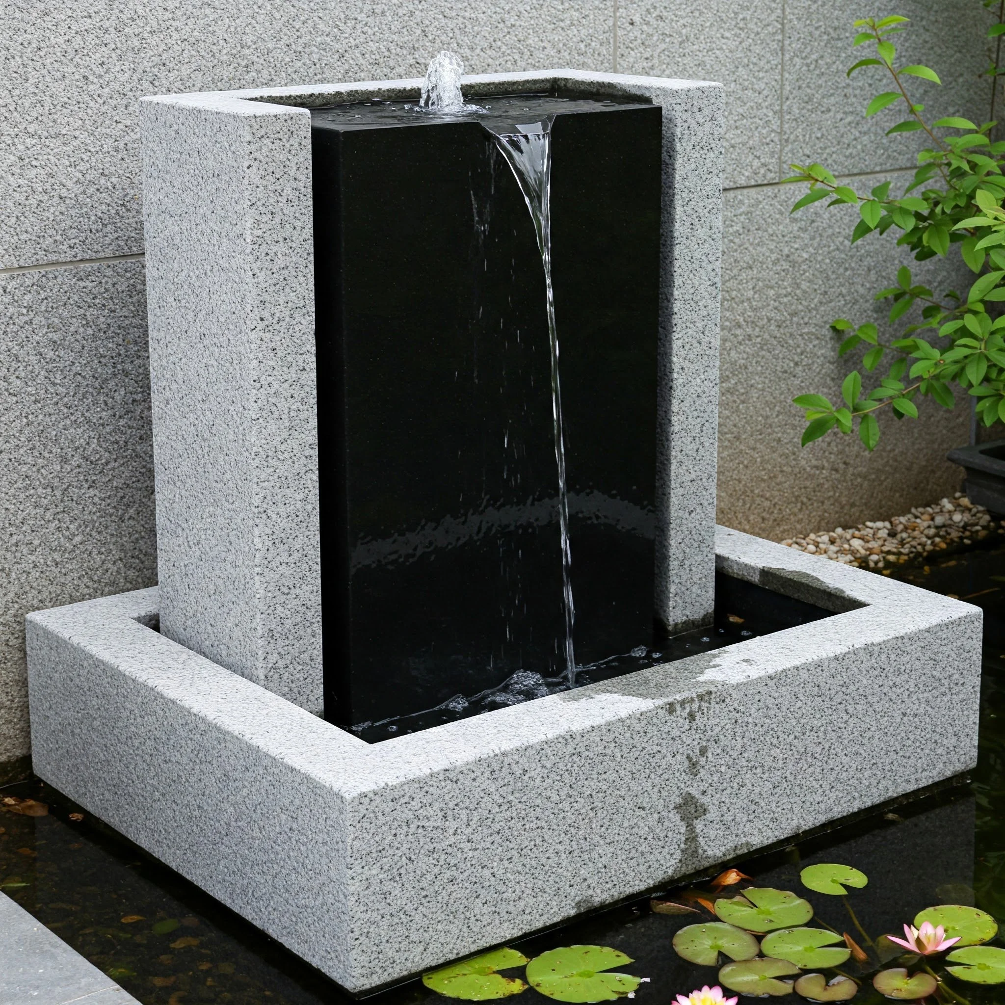 A modern outdoor water fountain with a rectangular stone structure and water flowing from a spout into a black basin, surrounded by lily pads and green plants.