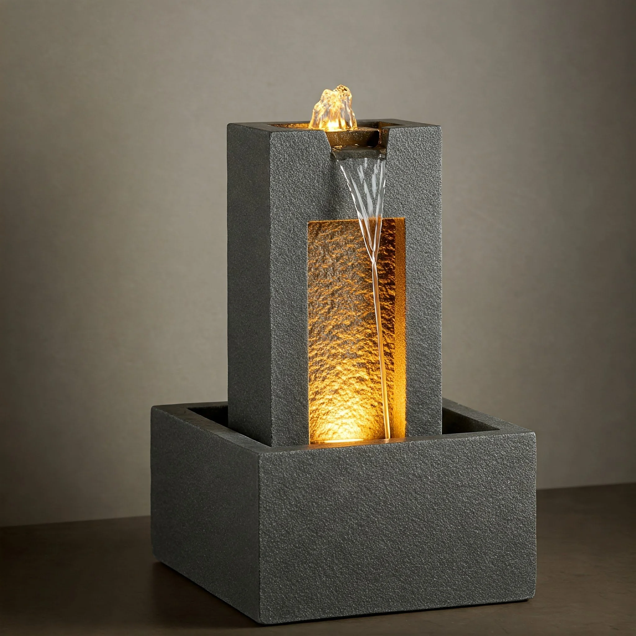 A modern tabletop water fountain with a rectangular base and a textured, stone-like appearance. Water flows from a small, sculpted top feature into a vertical basin below, illuminated by warm internal lighting. The background is plain and neutral, hi