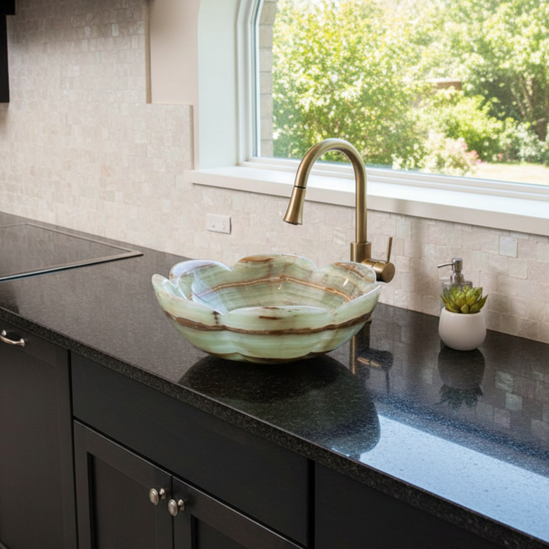 A kitchen countertop with a black surface, a decorative shell-shaped sink, a gold faucet, a small potted succulent, and a window with a view of green trees outside.