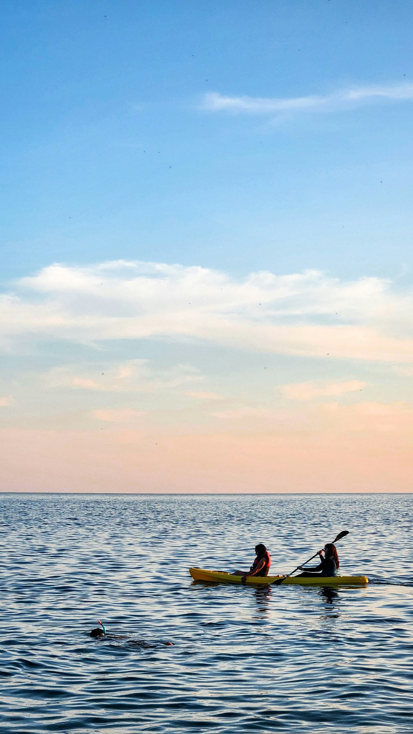 Two people kayaking on calm water during sunset with a swimmer nearby, under a pastel-colored sky.