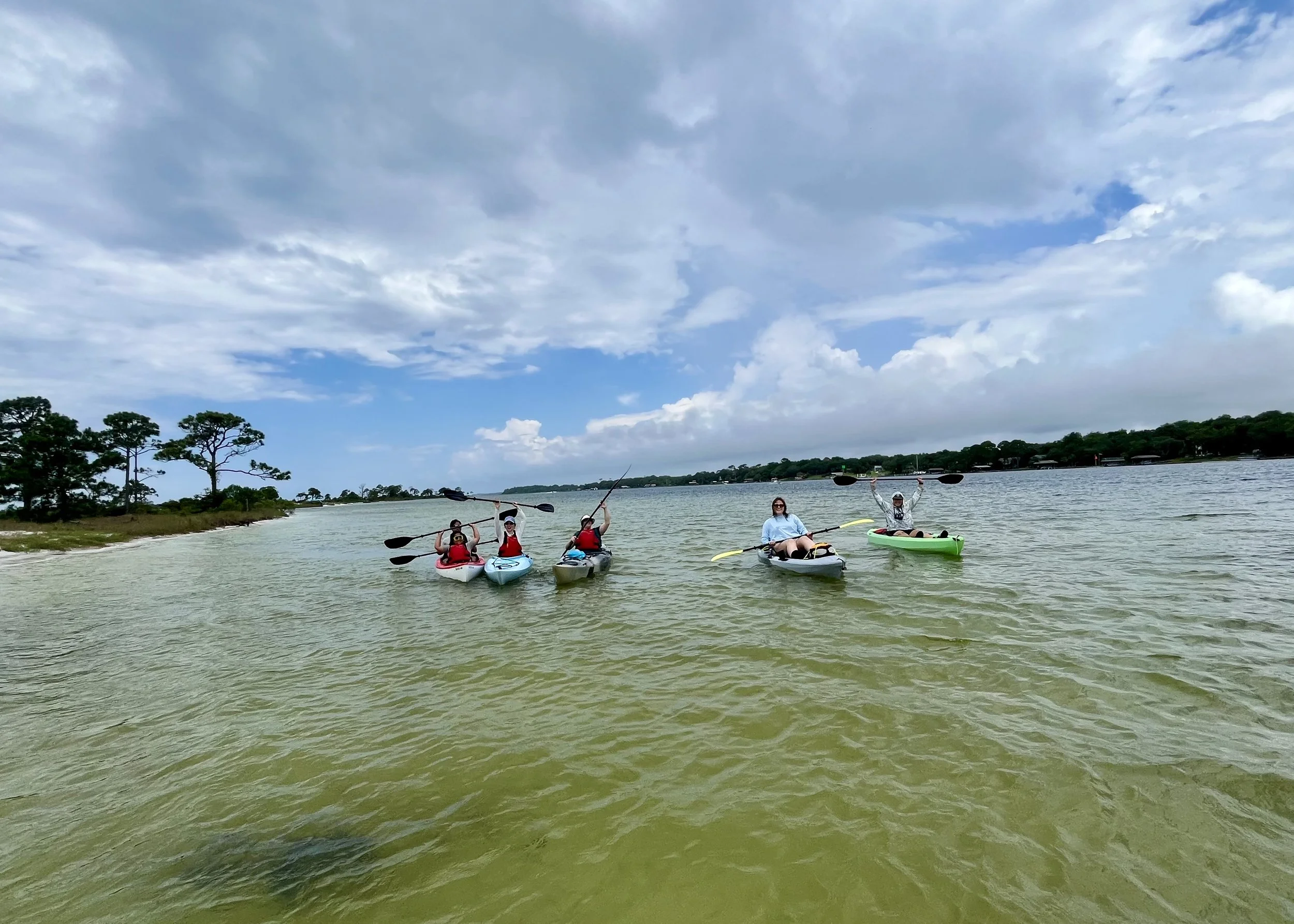 Four people kayaking on a lake with trees and a cloudy sky in the background.