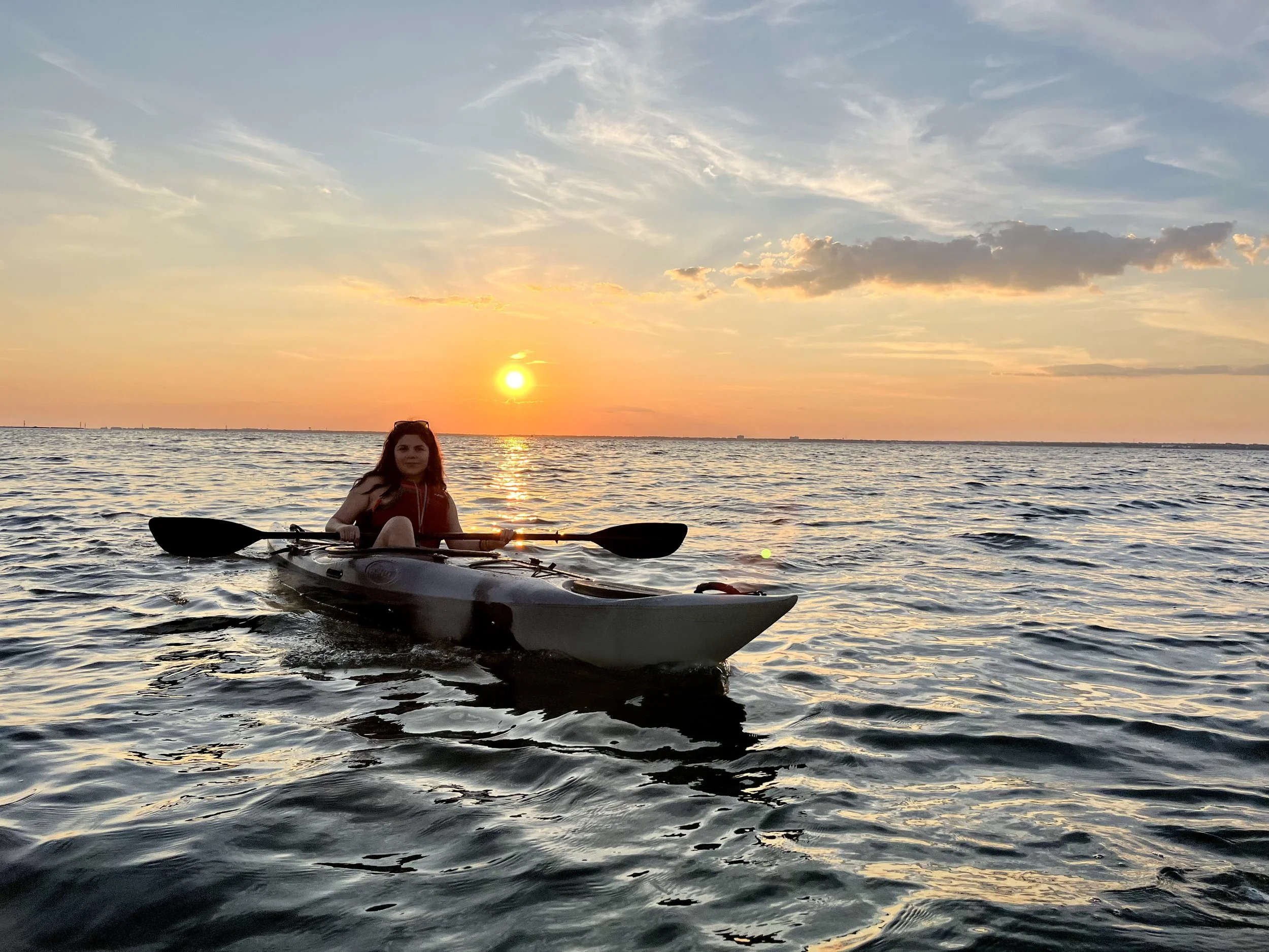 A woman kayaking on a body of water during sunset with a colorful sky and clouds.