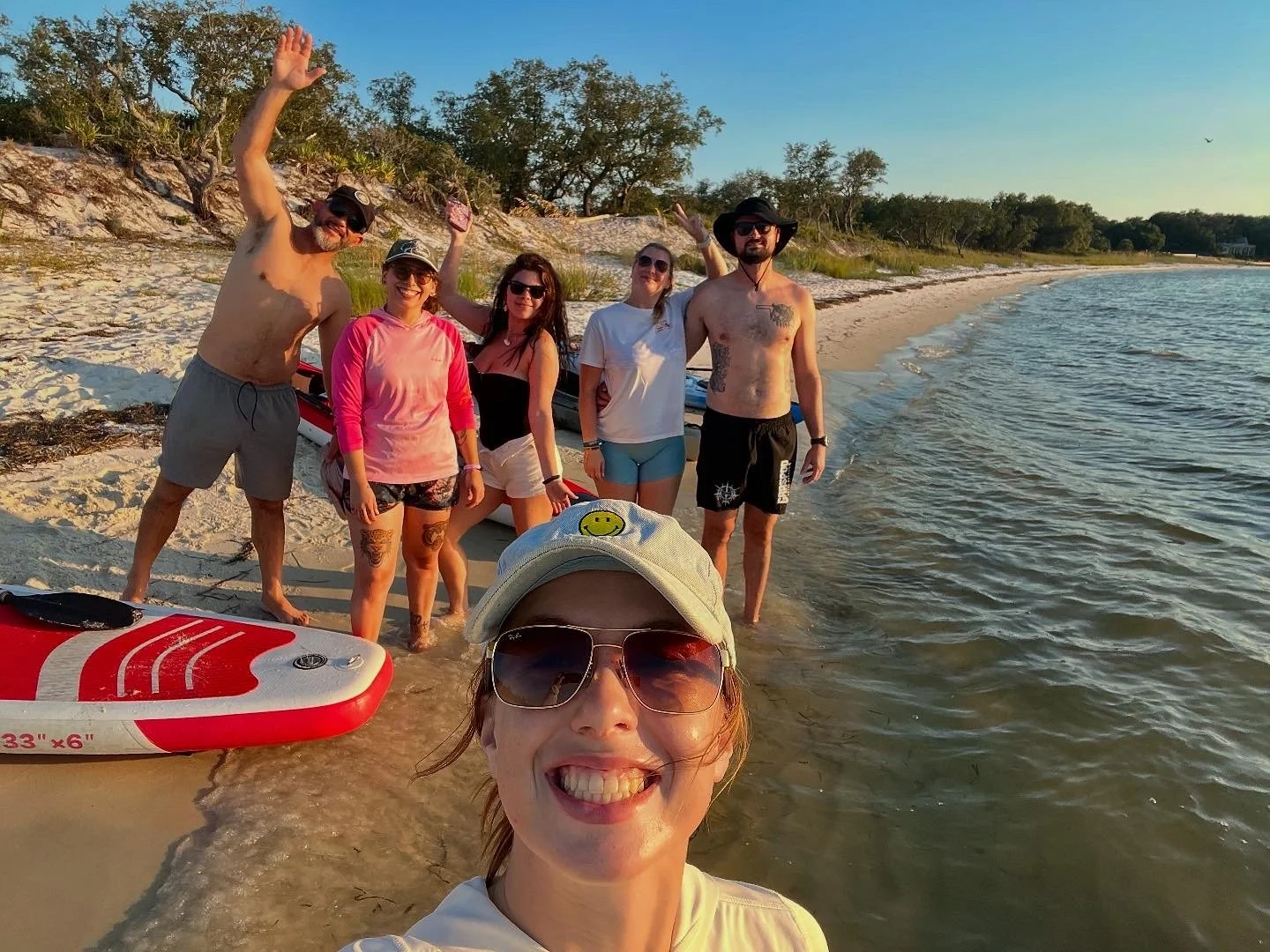 A group of six people on a beach, posing for a photo with a woman taking a selfie in the foreground, with some people standing in the shallow water and others on the sand, along with a paddleboard nearby, trees in the background, and a clear sky.