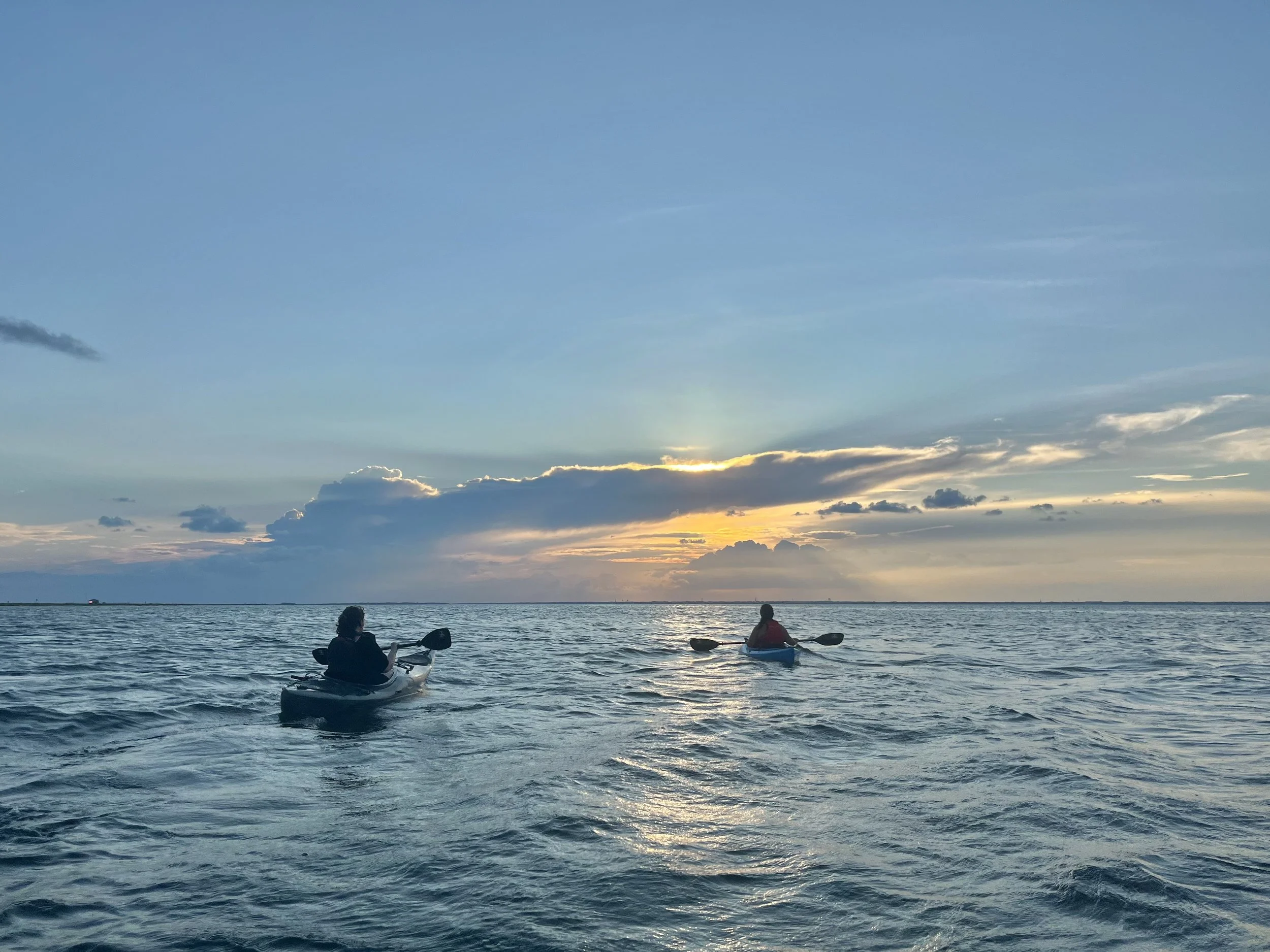 Two people kayaking on the ocean during sunset with partly cloudy sky.