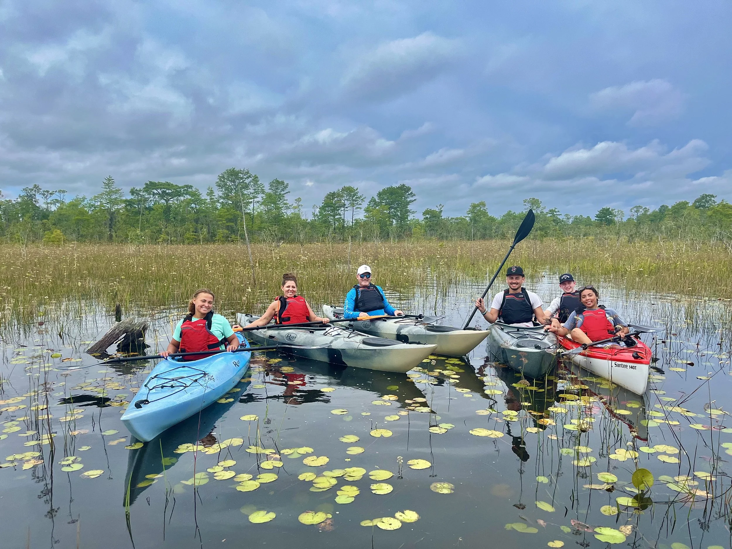 Group of five people kayaking on a calm marsh with water lilies, tall grass, and trees in the background under cloudy sky.