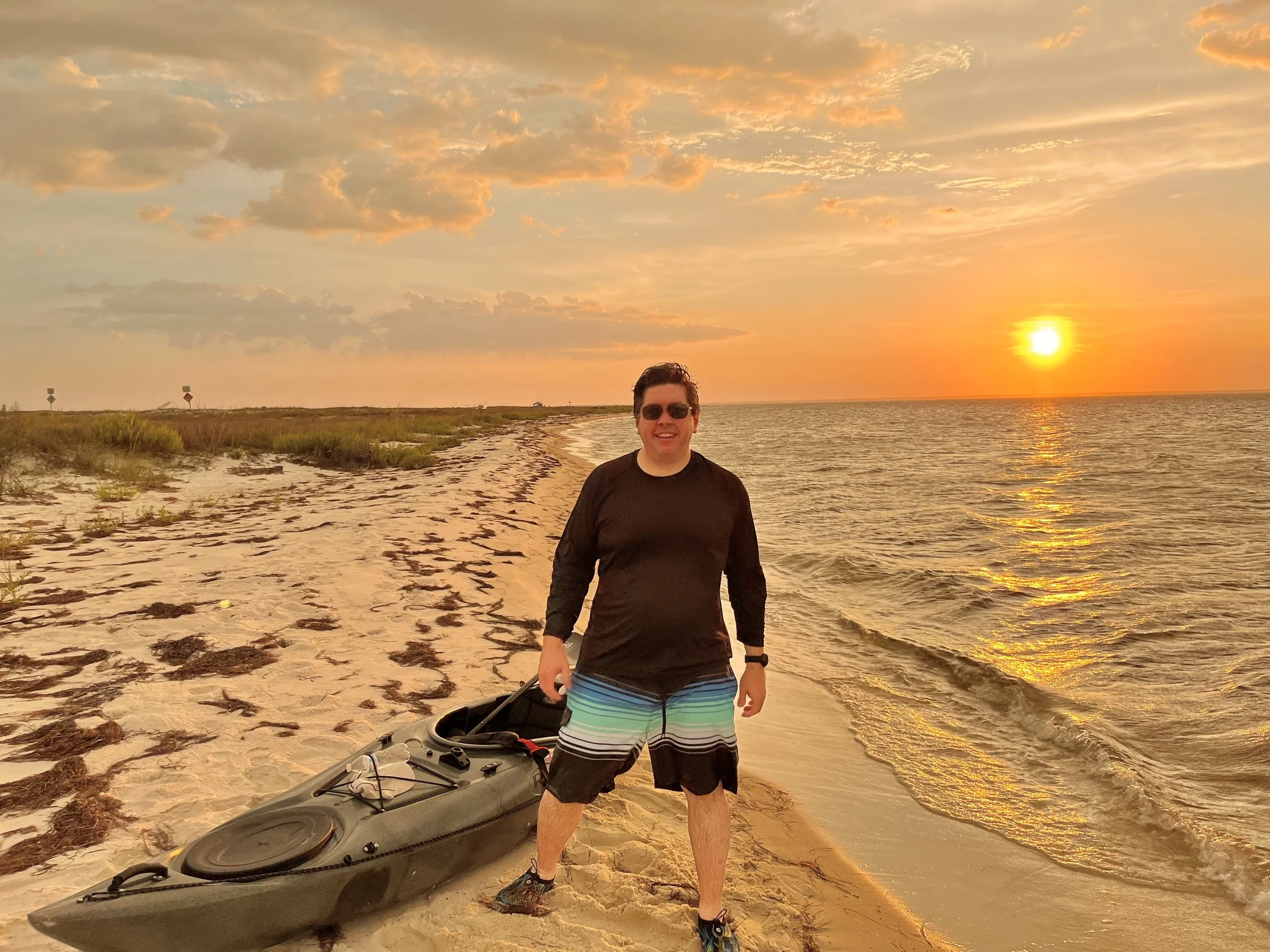 A man wearing sunglasses, a black long-sleeve shirt, and striped swim trunks stands on a beach near a kayak during a sunset, with the sun reflecting on the water.