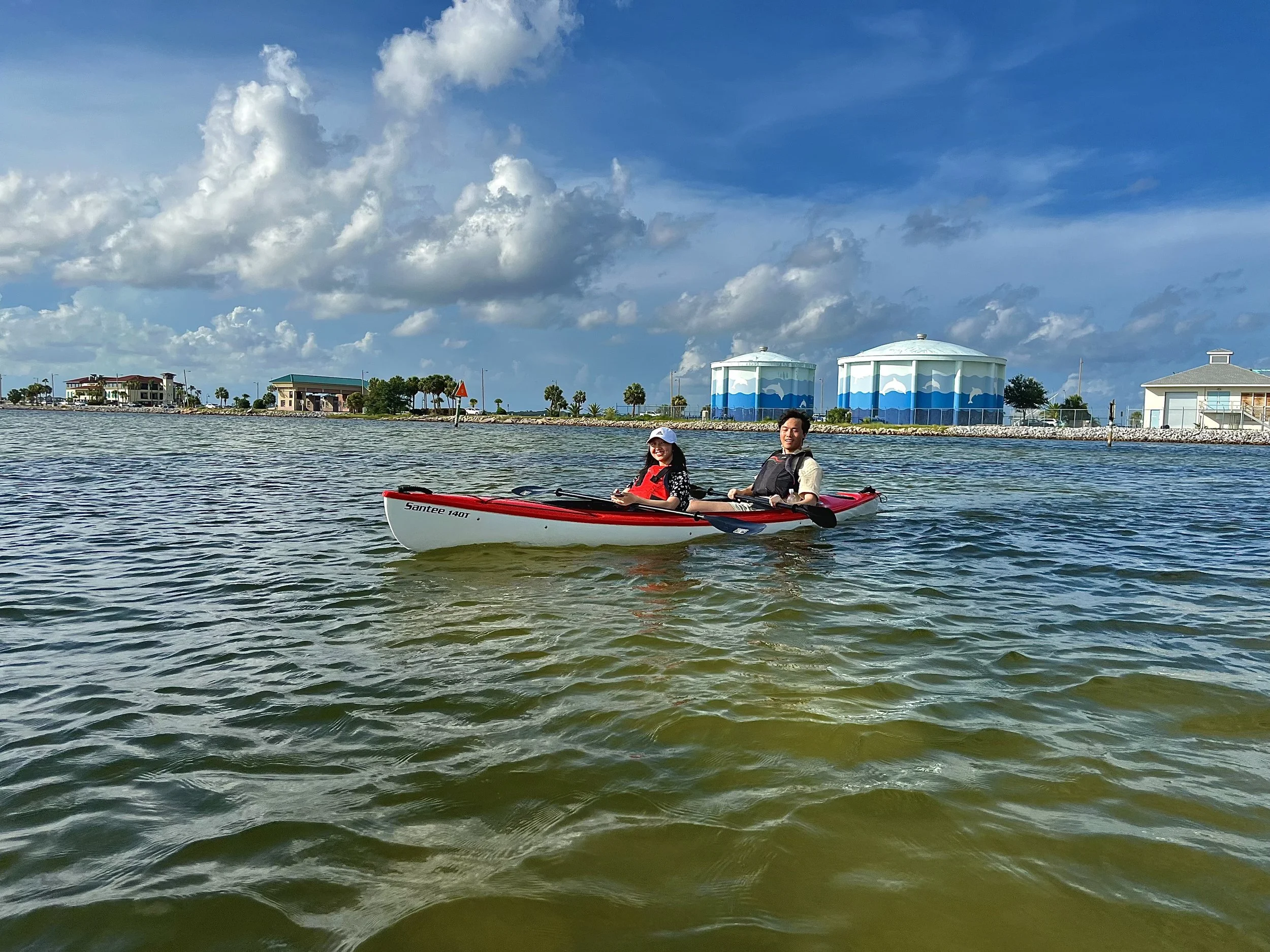 Two people kayaking on a body of water with a cloudy sky and buildings in the background.