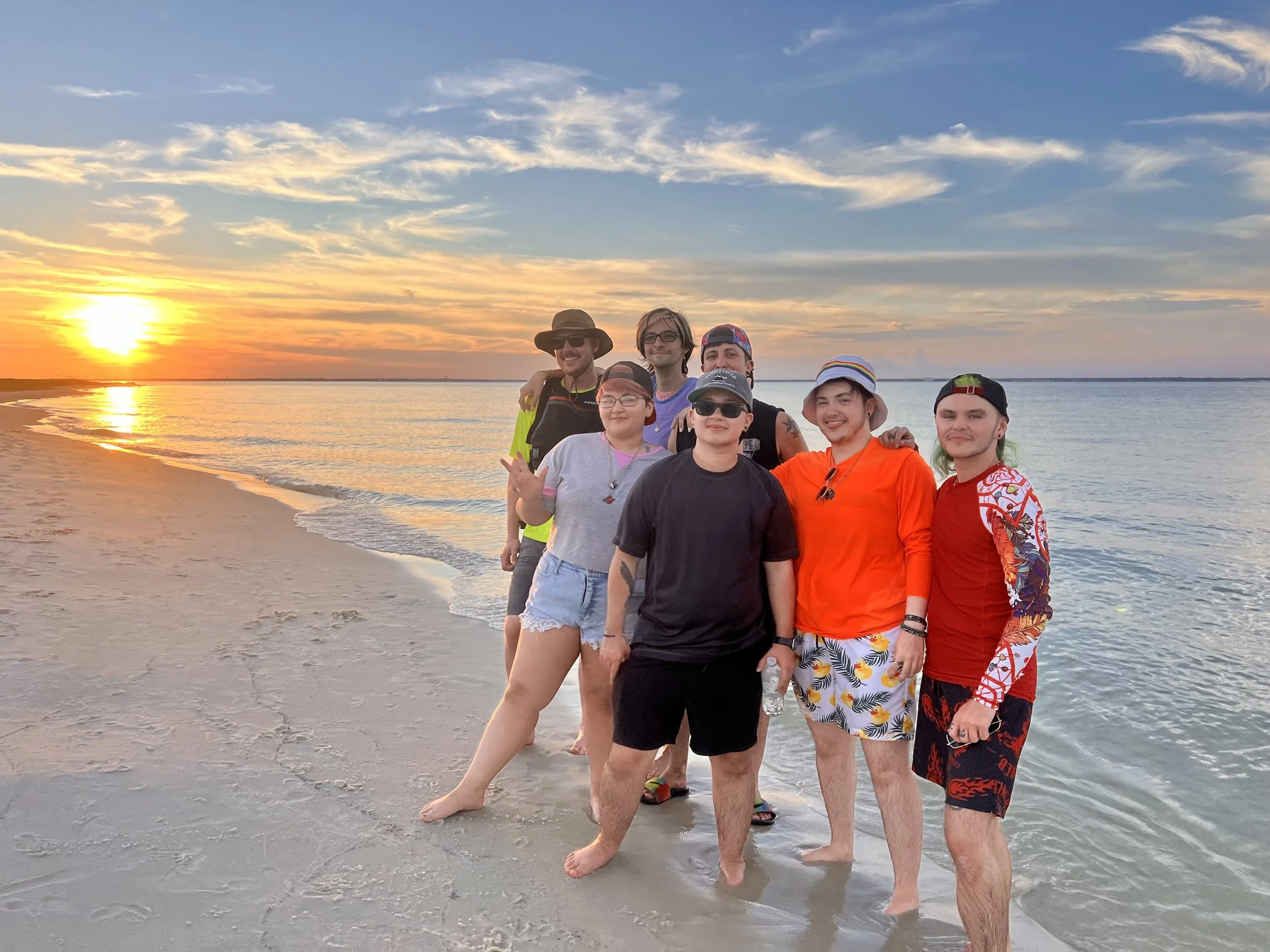 Group of seven young adults standing on a beach at sunset, dressed in summer casuals, with the water and sky in the background.