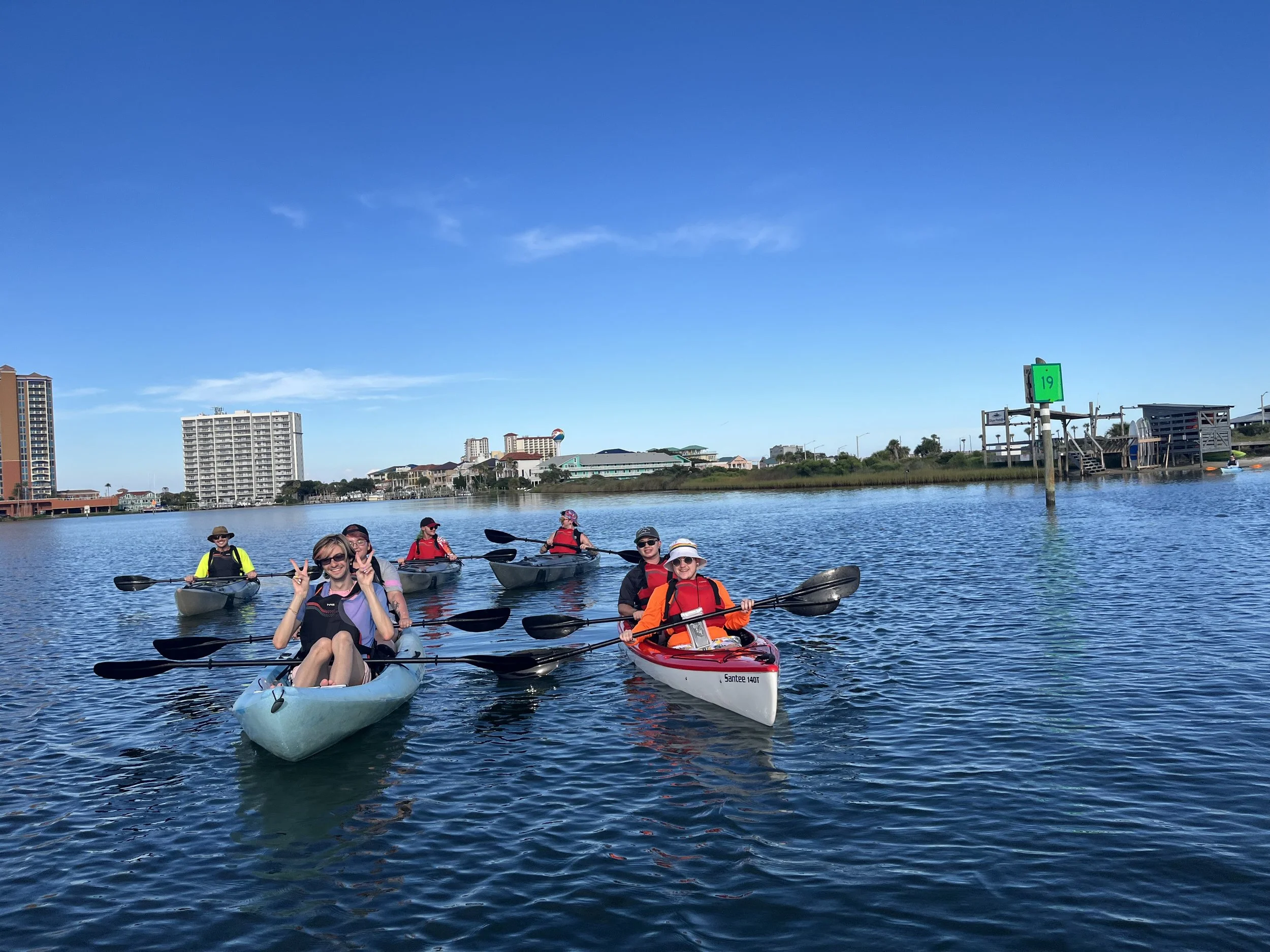 Group of six people kayaking on a calm body of water on a sunny day, with city buildings and a dock in the background.