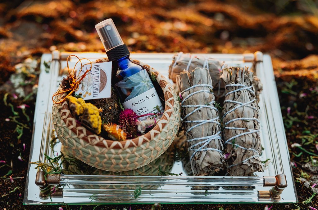 A decorated tray displaying a woven basket with dried flowers and a blue spray bottle labeled 'White Sage.' Next to the basket are three bundles of sage tied with string.