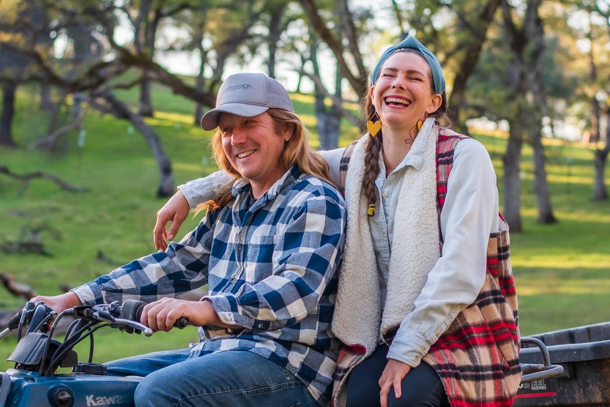 A man in a checkered shirt and cap sits on a motorcycle with a smiling woman, wearing a plaid vest, leaning against him. They are outdoors in a grassy area with trees in the background.