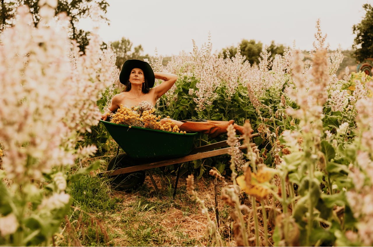 A person wearing a hat relaxing in a wheelbarrow filled with flowers in a lush garden.