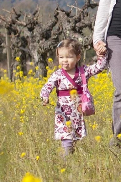 Young girl in a pink floral kimono with a small pink purse, holding hands with an adult, walking through a field of yellow flowers with trees in the background.