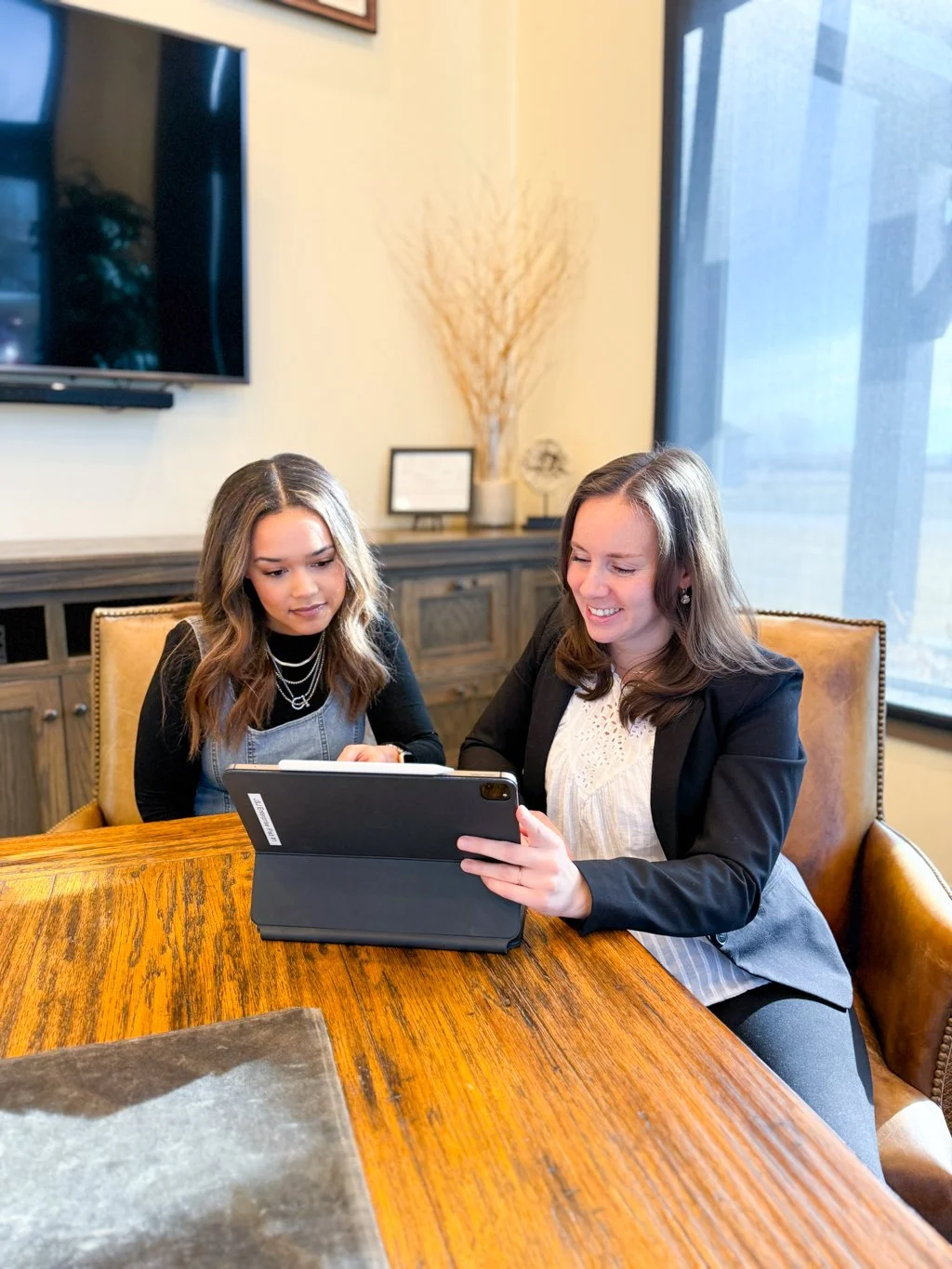 Two beautiful warm welcoming women sitting at a wooden table in an office setting looking at a tablet. A window and office furniture are visible in the background. Warm, welcoming, and inviting.