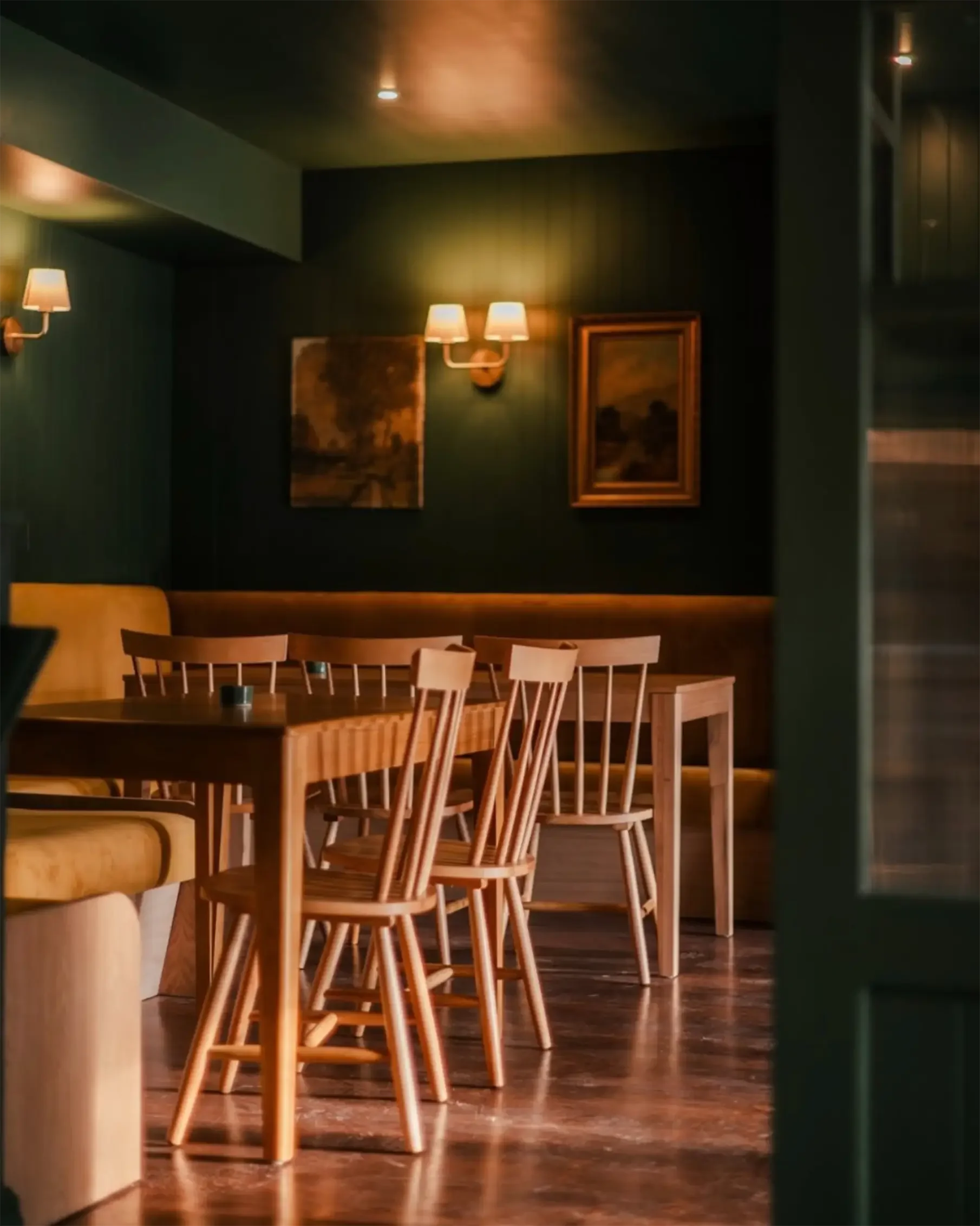 The Captain Darling, Stockbridge interior, empty dining area with wooden table and chairs, dark wall with framed paintings, and warm wall-mounted lights in a cosy restaurant.