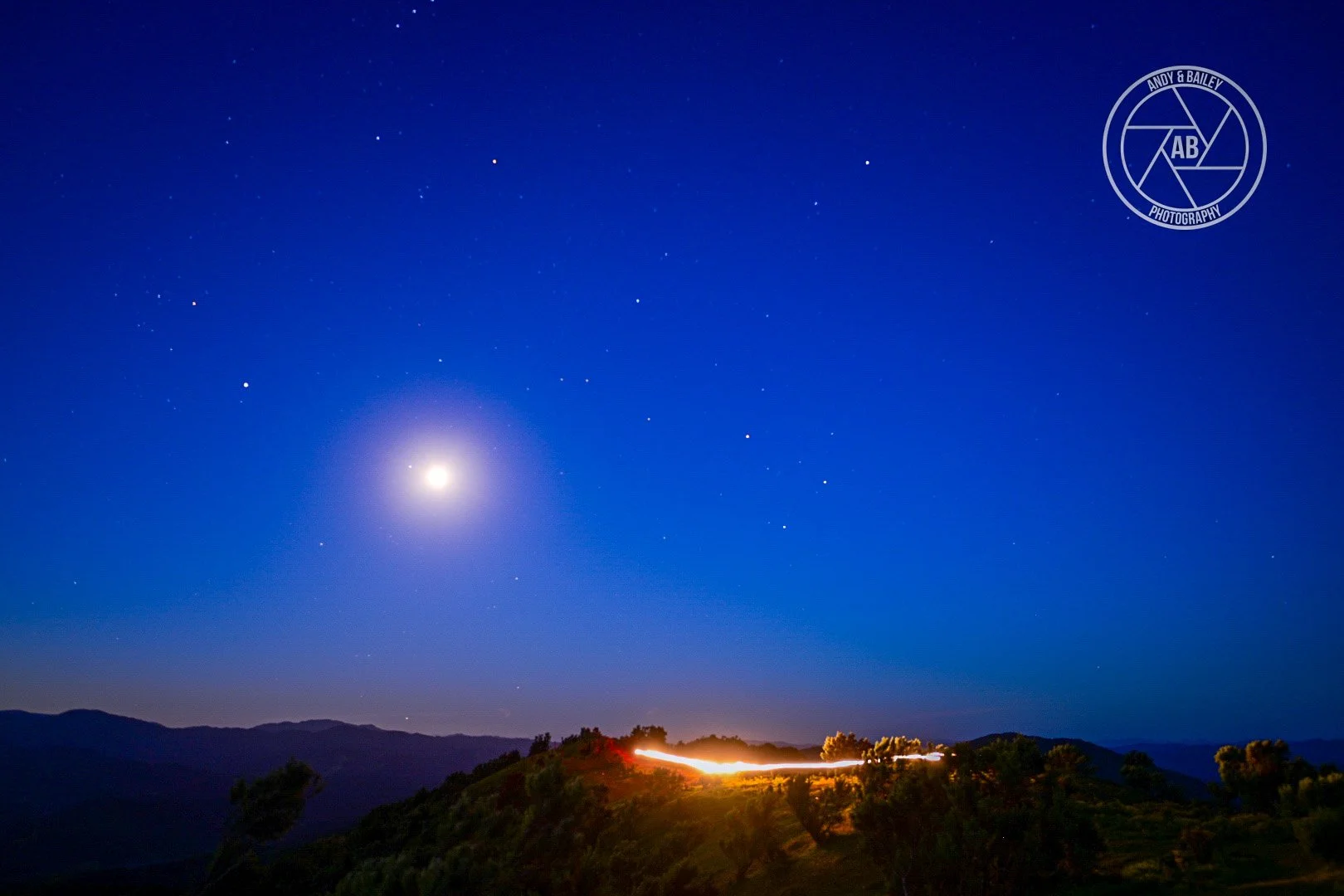 Nighttime landscape with moon, stars, and a distant trail of light over hills