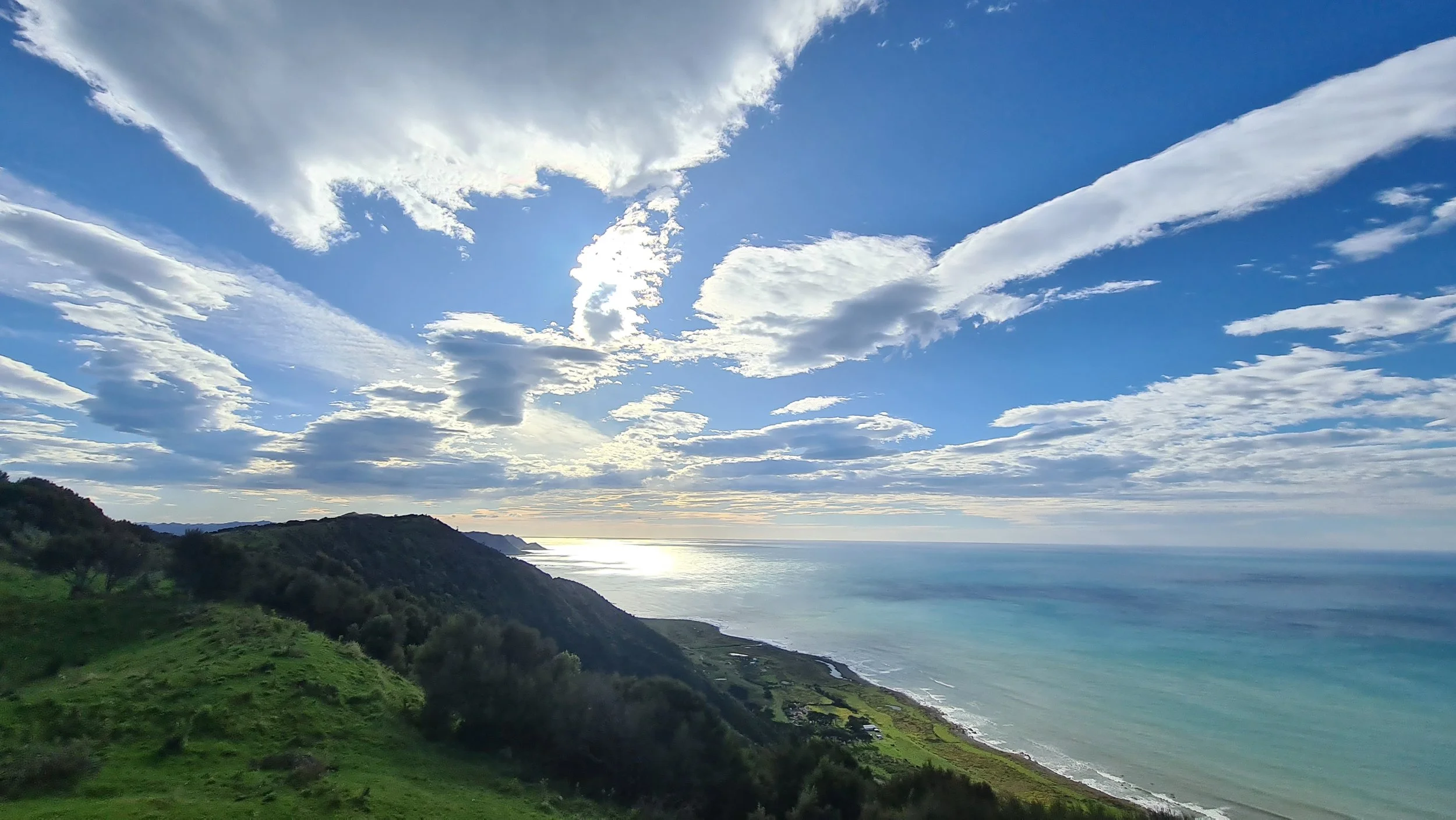 A scenic view of a coastal landscape with green hills, a calm ocean, and a blue sky filled with white clouds.