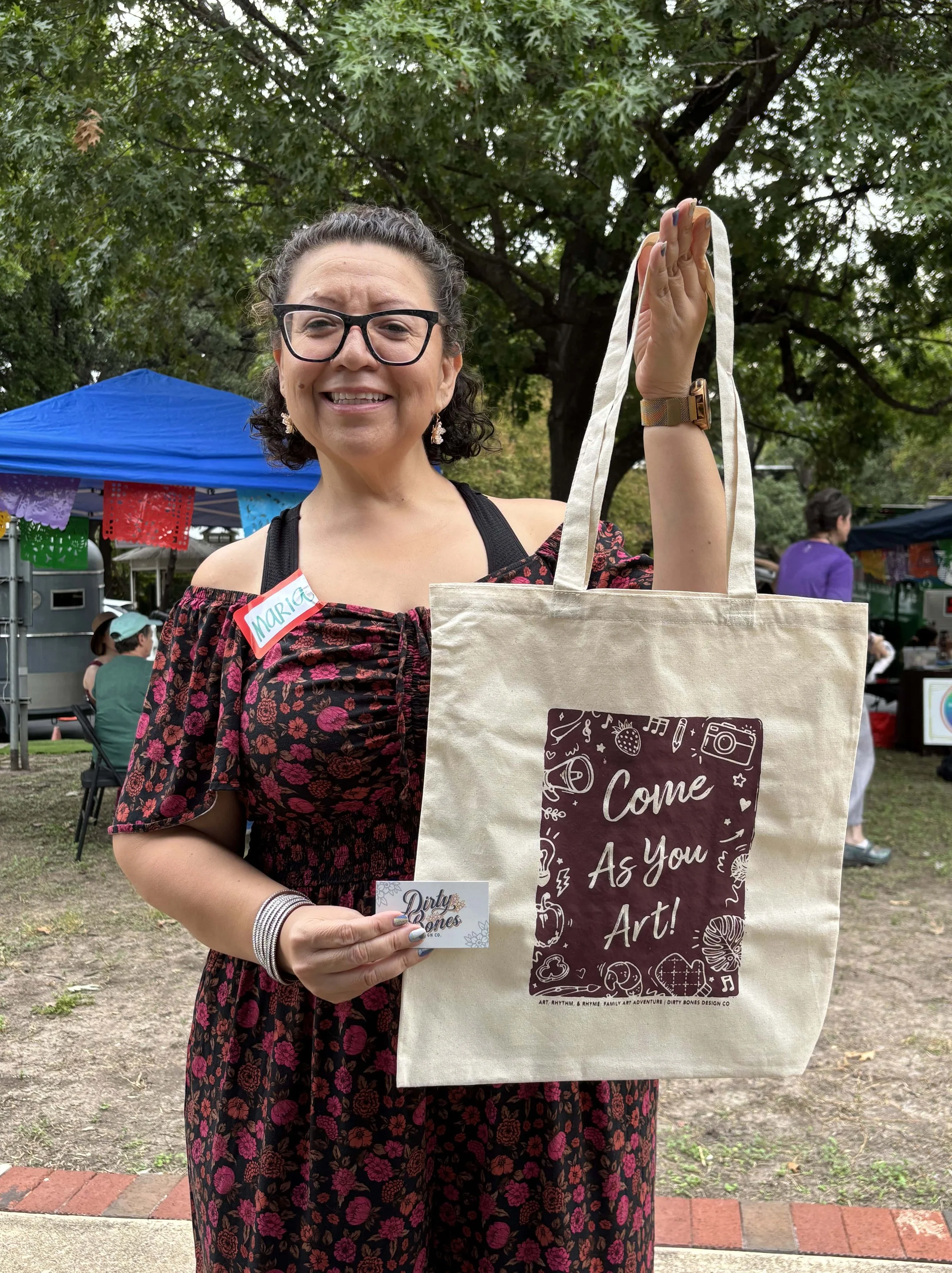 A woman with glasses and curly hair standing outdoors at a fair or market, holding a tote bag with the message 'Come As You Art!' and a business card that says 'Dirty Bones'. She is wearing a black dress with a floral pattern, has a name tag reading 'Maria', and is smiling.