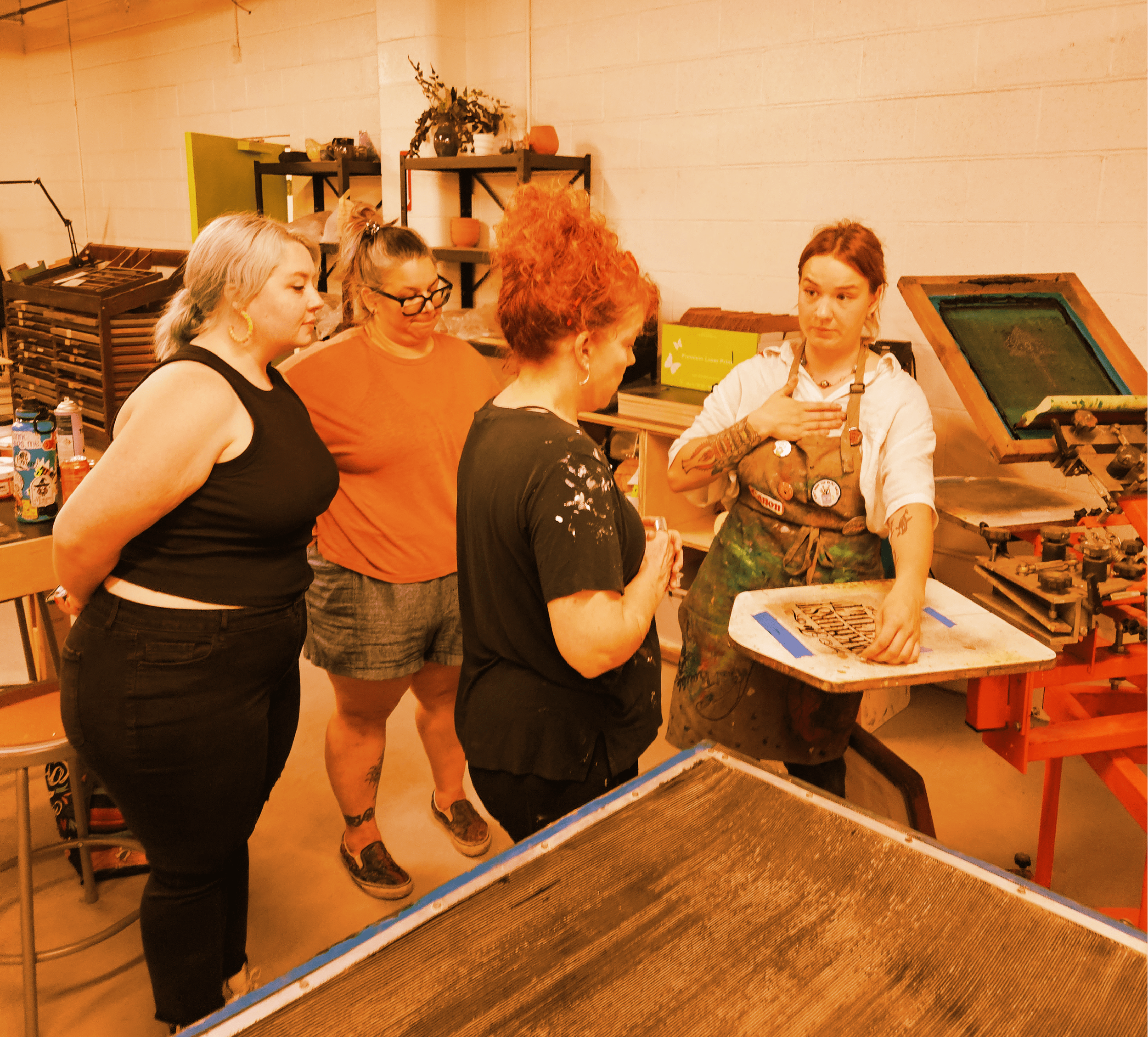 Four women in an art workshop, two observing and two demonstrating printmaking techniques using an etching press.