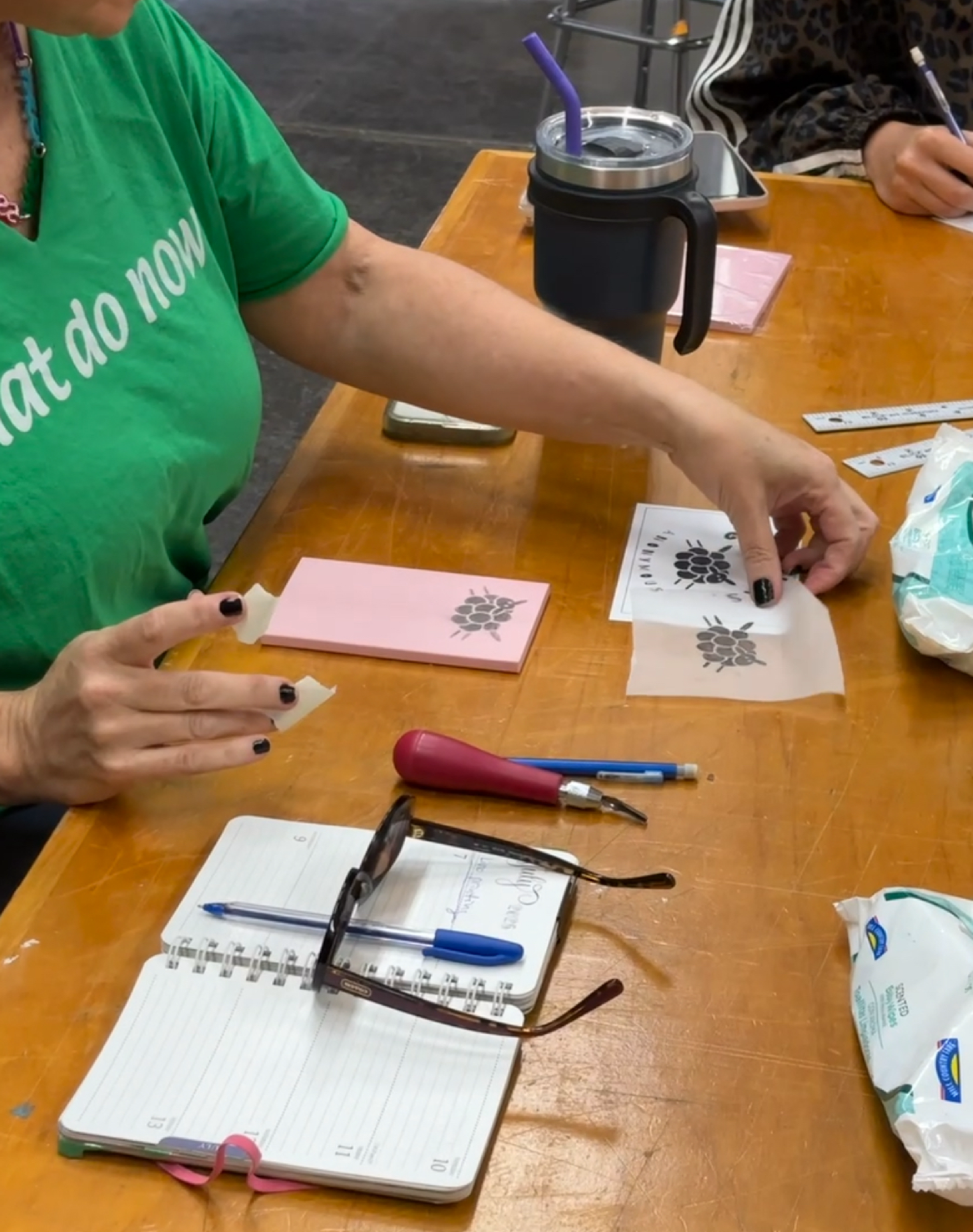 Person sitting at a wooden table, applying a tattoo stencil onto paper. Various tools, notebooks, and a drink with a purple straw are on the table.