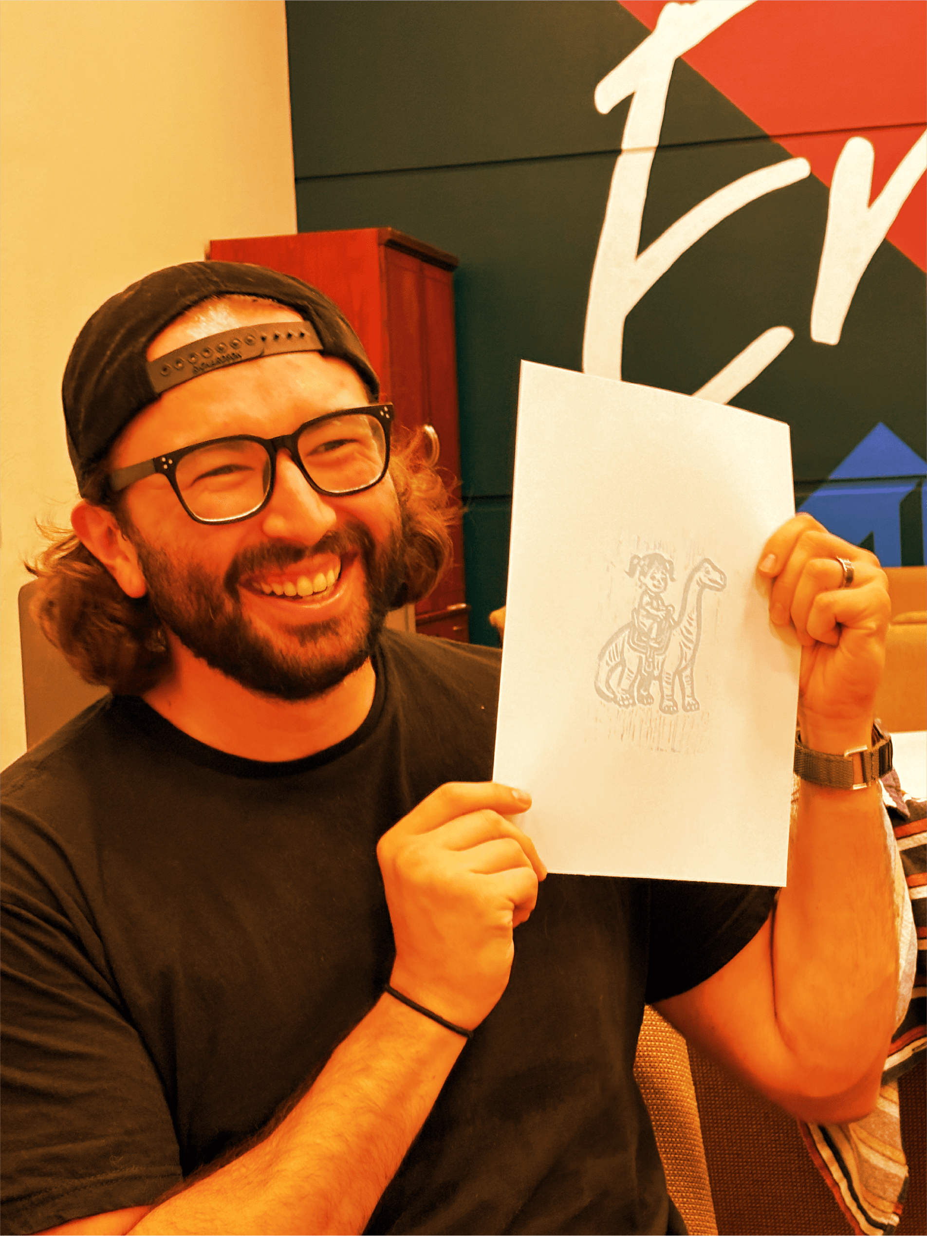 A smiling man with glasses, a black T-shirt, and a backward cap holding a drawing of a person riding a dinosaur.