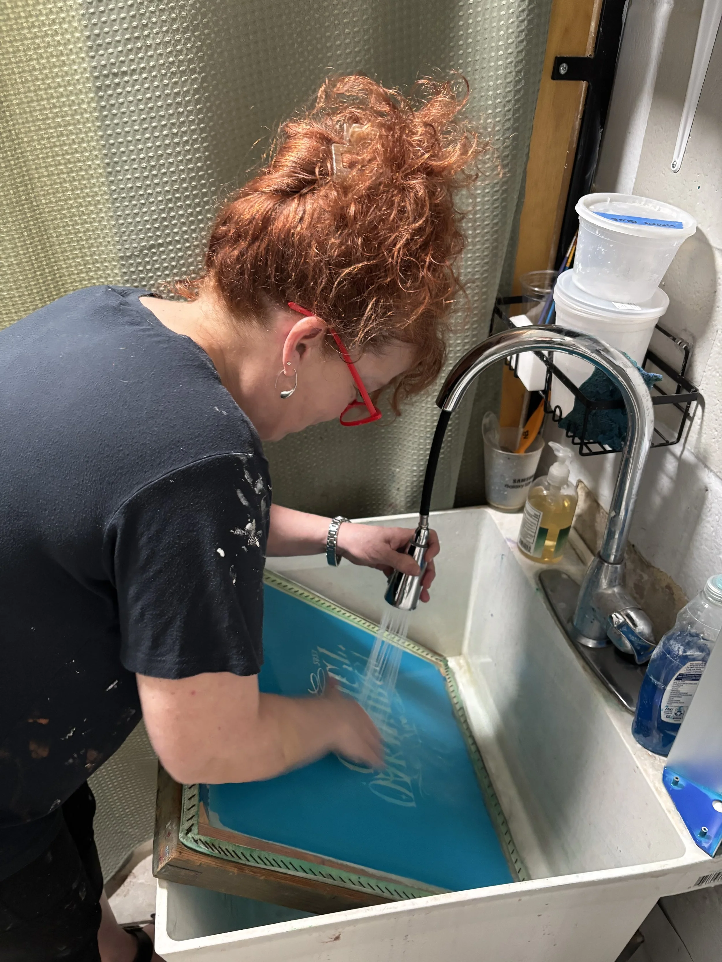 Woman with red hair and glasses washing a blue screen printing frame in a sink.