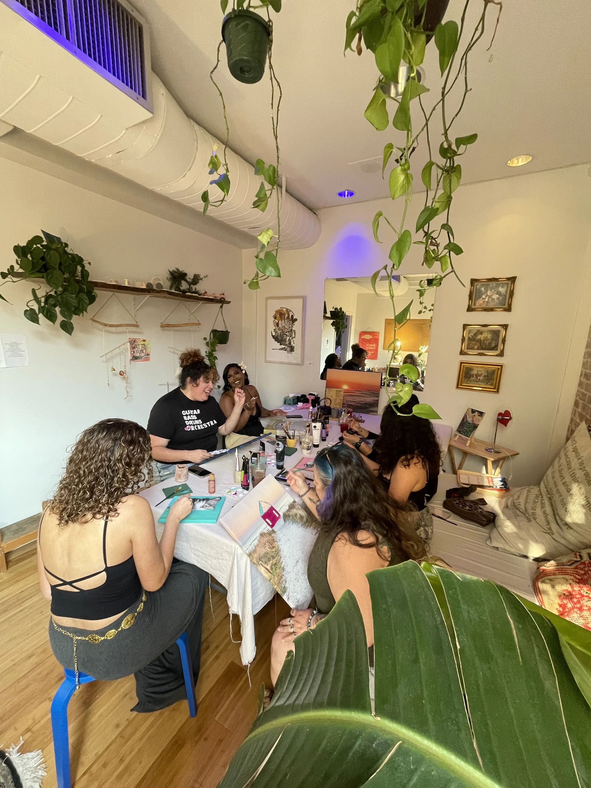 A group of women sitting around a table engaged in a craft or makeup activity in a cozy, decorated room with hanging plants, framed artwork, and a large green leaf in the foreground.