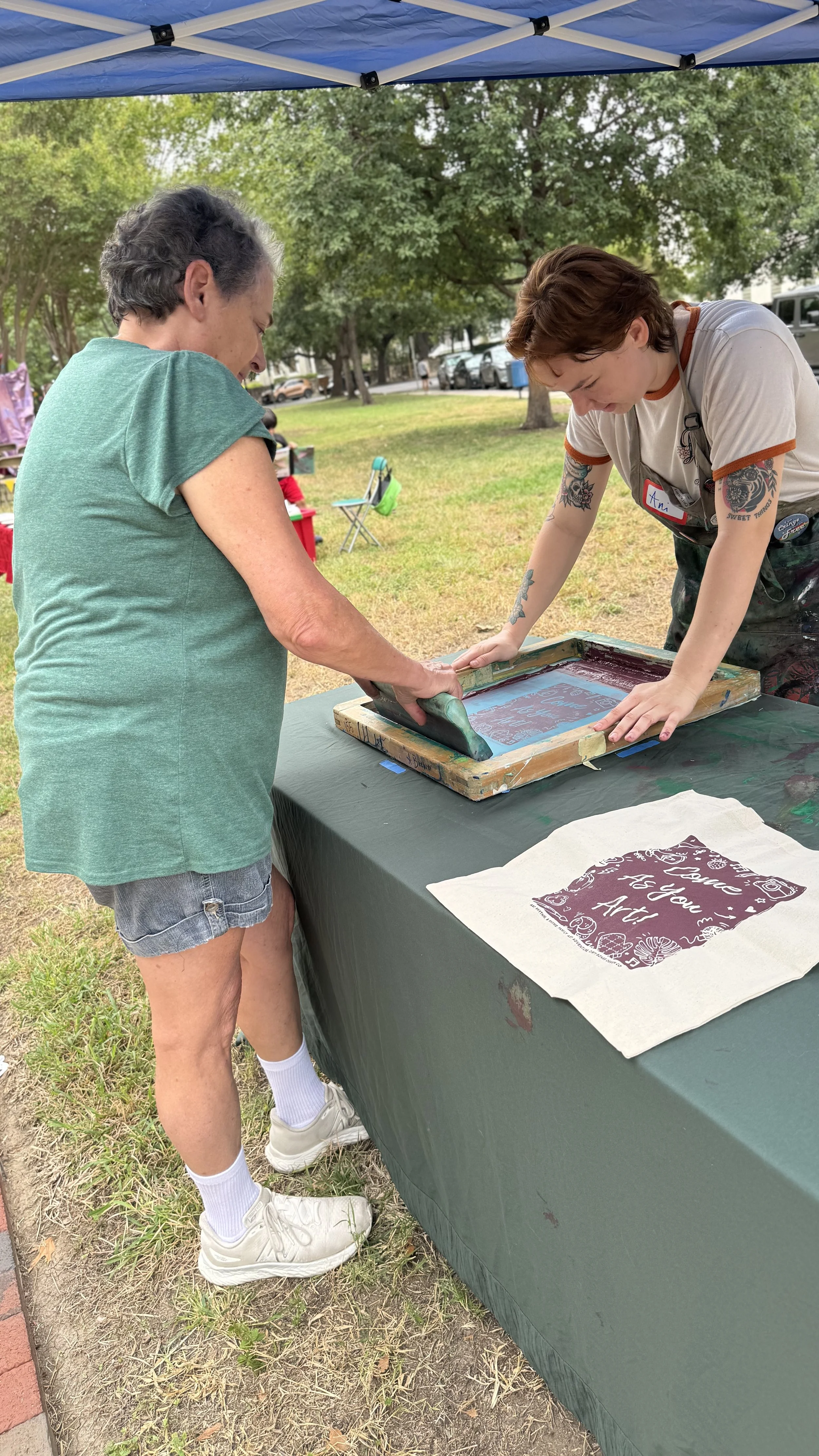Two women are participating in a screen printing activity at an outdoor event, with a blue canopy overhead and green trees in the background.