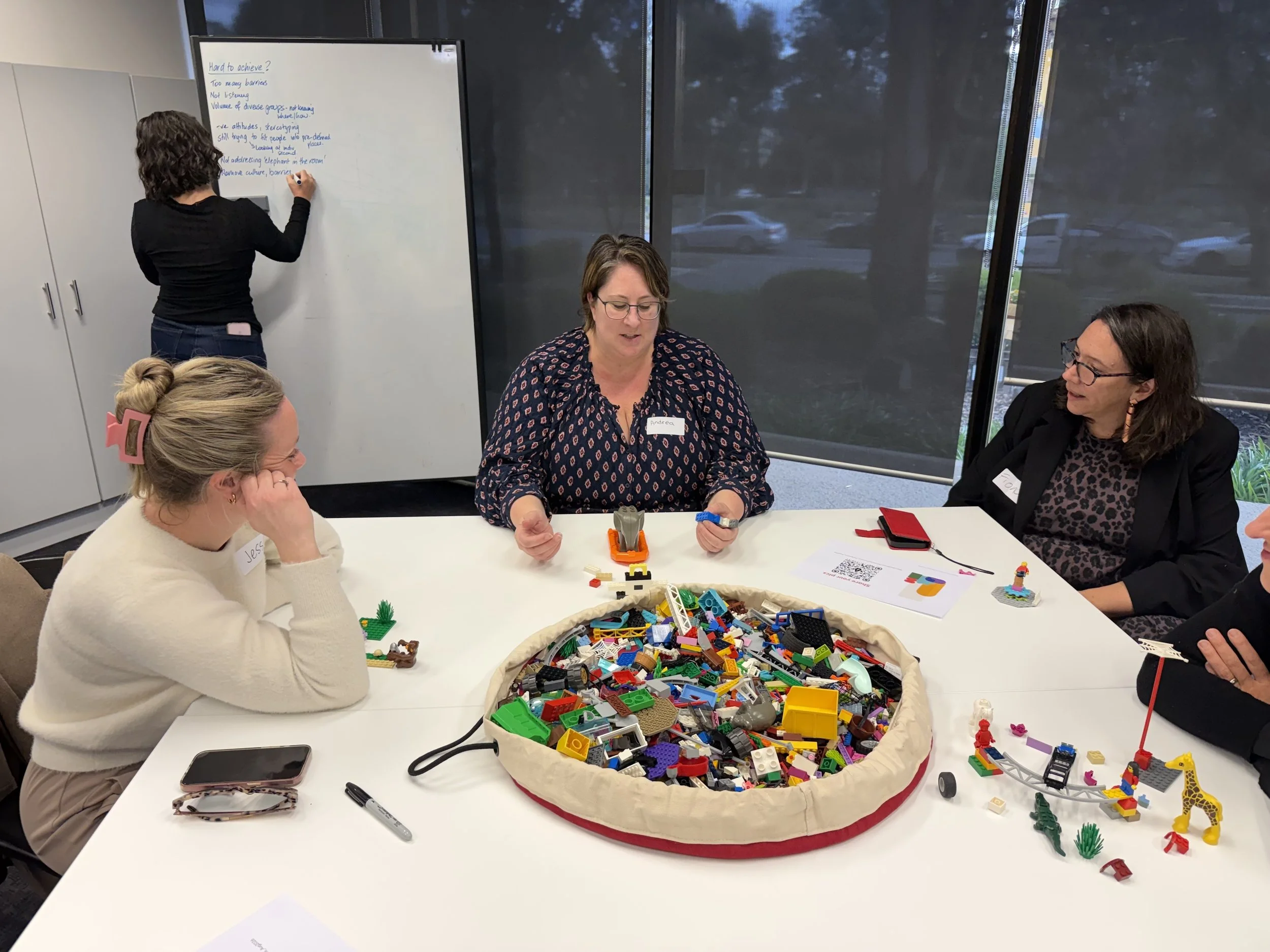Three people discussing a LEGO model build with someone writing notes on a whiteboard behind them