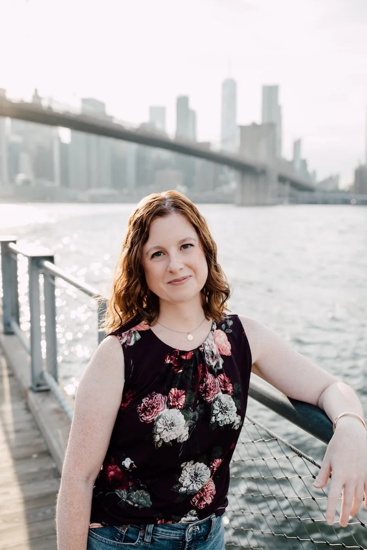 Jessica Plummer smiling at the camera, with the East River and Brooklyn Bridge behind her.