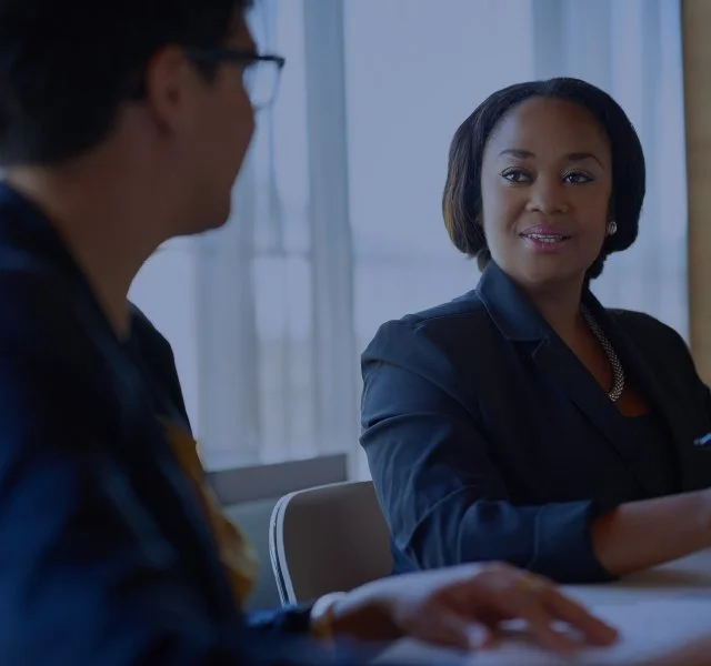 Two women in professional attire having a conversation in an office setting with large windows.