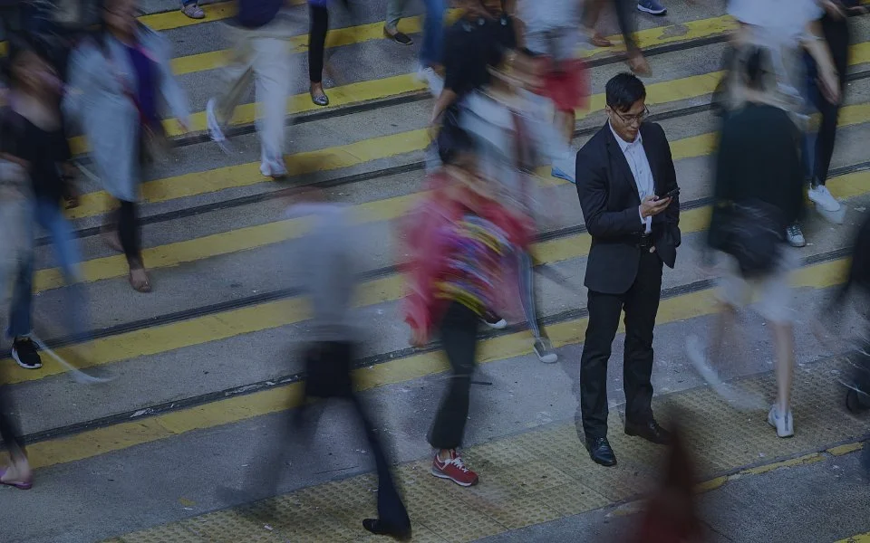 A busy crosswalk with many people walking, some blurred from motion. One man in a suit stands still looking at his phone amidst the crowd.