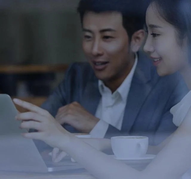 Two people sit at a table, looking at a tablet together, with a cup on the table, in a dimly lit setting.