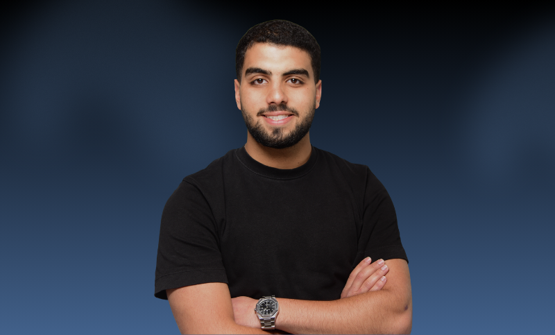 A young man with short dark hair and a beard, wearing a black t-shirt and a silver watch, standing with his arms crossed and smiling against a dark blue background.