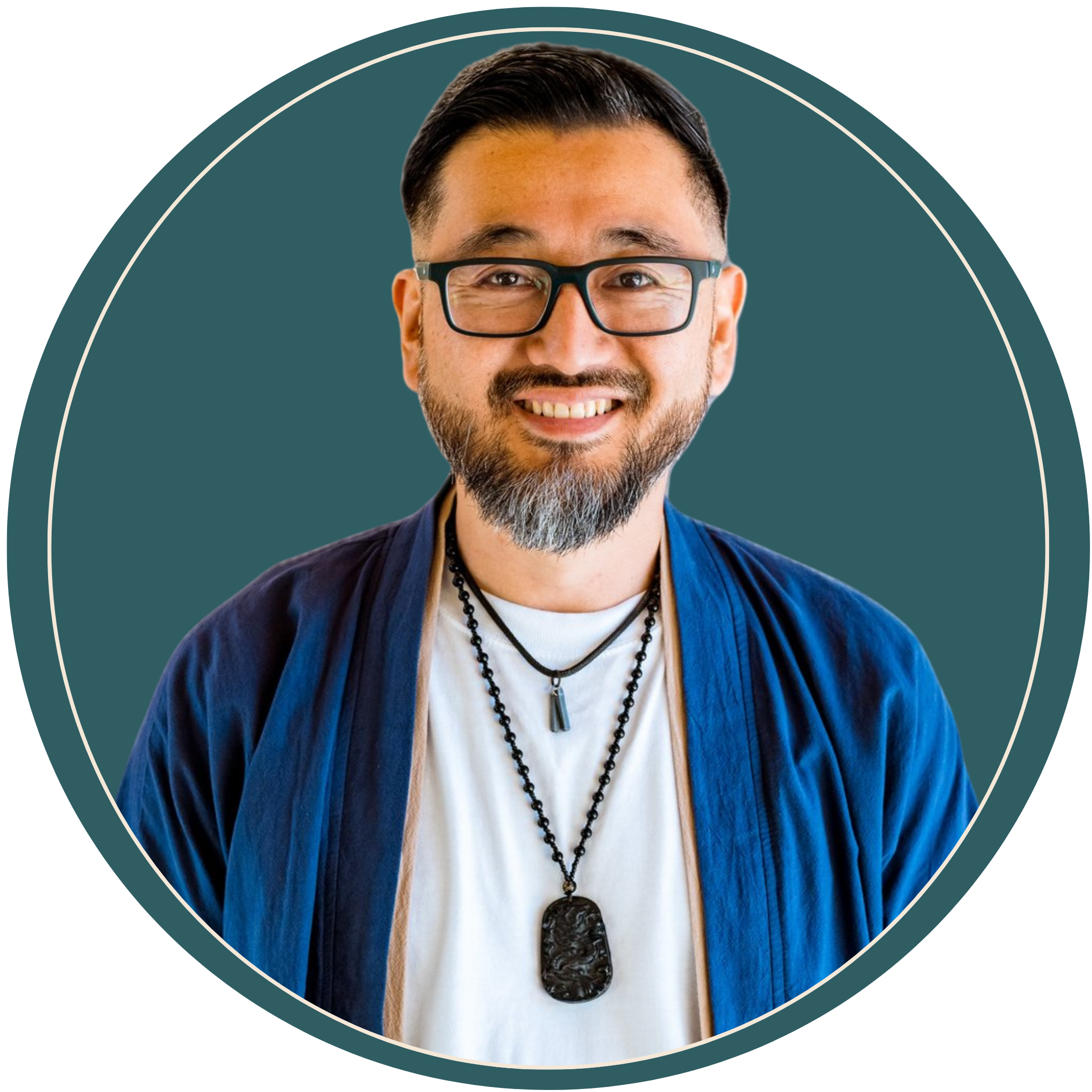 Smiling man with glasses and a beard wearing a white shirt and a blue jacket, with layered necklaces, against a teal background.