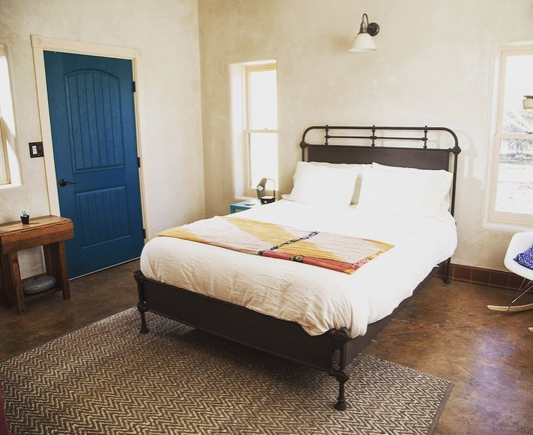 A bedroom with a black metal bed frame, white bedding, two pillows, and a colorful throw blanket. There are two windows beside the bed, a small blue nightstand with an alarm clock, a wall-mounted lamp overhead, a wooden side table with a small plant,