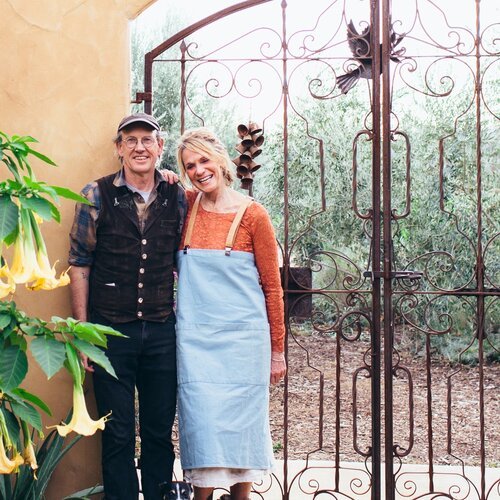 A smiling man and woman standing together in front of a decorative metal gate and garden, with yellow flowers on the left.