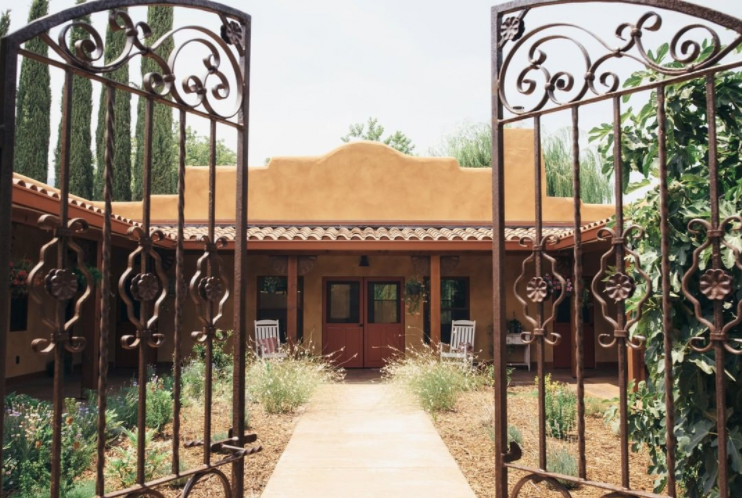 View through decorative wrought iron gate leading to a southwestern style house with red doors, surrounded by garden and trees.