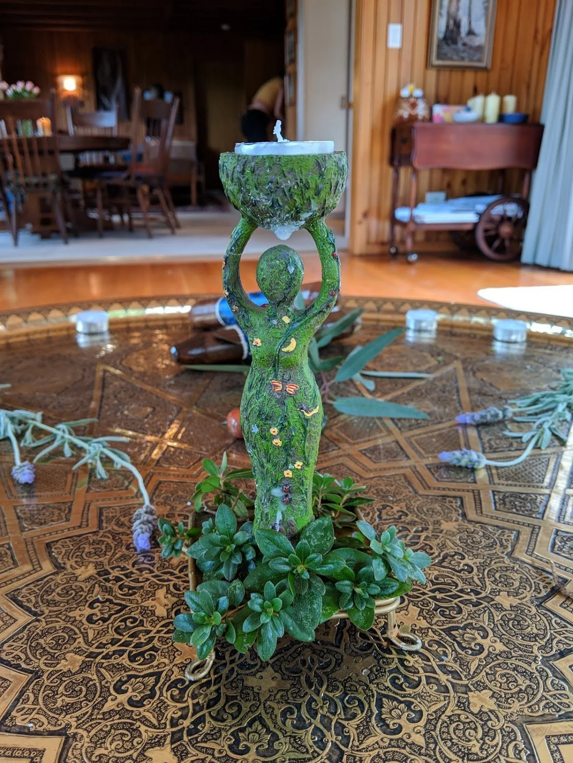 Decorative candle holder shaped like a woman, painted with floral designs, placed on a patterned wooden table with surrounding greenery and flowers, in a cozy wooden interior.
