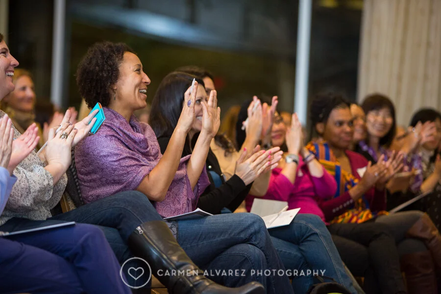 A group of diverse women seated indoors at a Wisdom 2.0 Women event, clapping and smiling during an event or presentation.