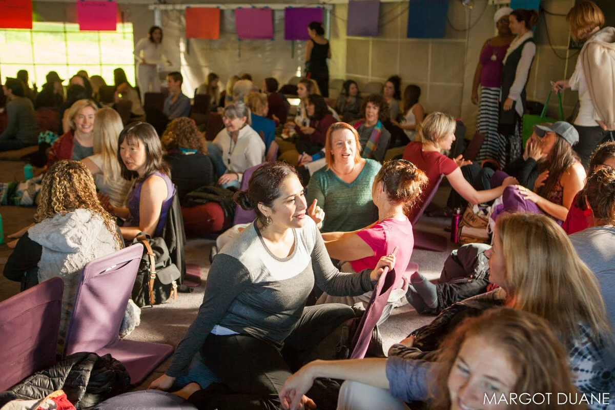 Women sitting on the floor and chairs inside a large room or hall, engaged in conversations, some smiling and laughing, with colorful decorations hanging above. Taken from a WisdomWomen Visionary Gathering at Esalen Institute.