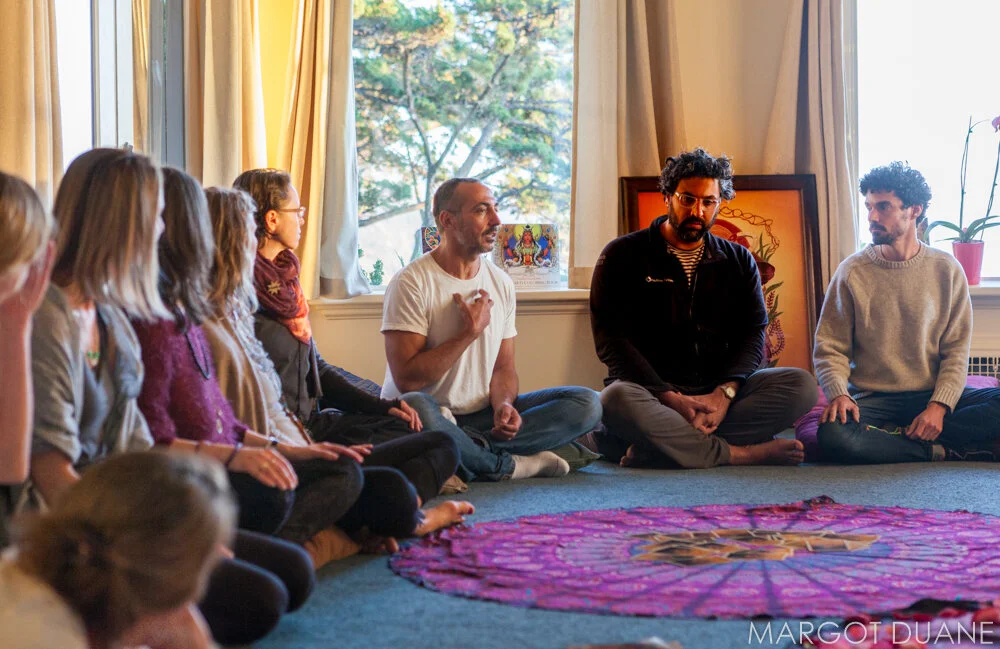 Group of people sitting cross-legged on the floor inside a room, engaged in a discussion or meditation, with a window showing trees in the background and colorful artwork behind them. This is from a WisodmWomen Visionary Gathering at Esalen Institute