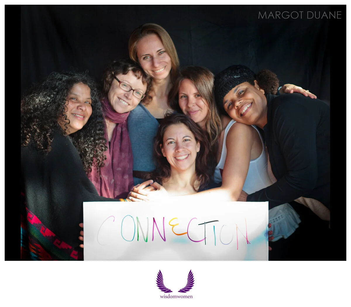 Group of six diverse women smiling and hugging around a sign that says 'CONNECTION' in colorful letters, against a black background. The photo is from WisdomWomen's Visionary Gathering at Esalen Institute in Big Sur, California.



