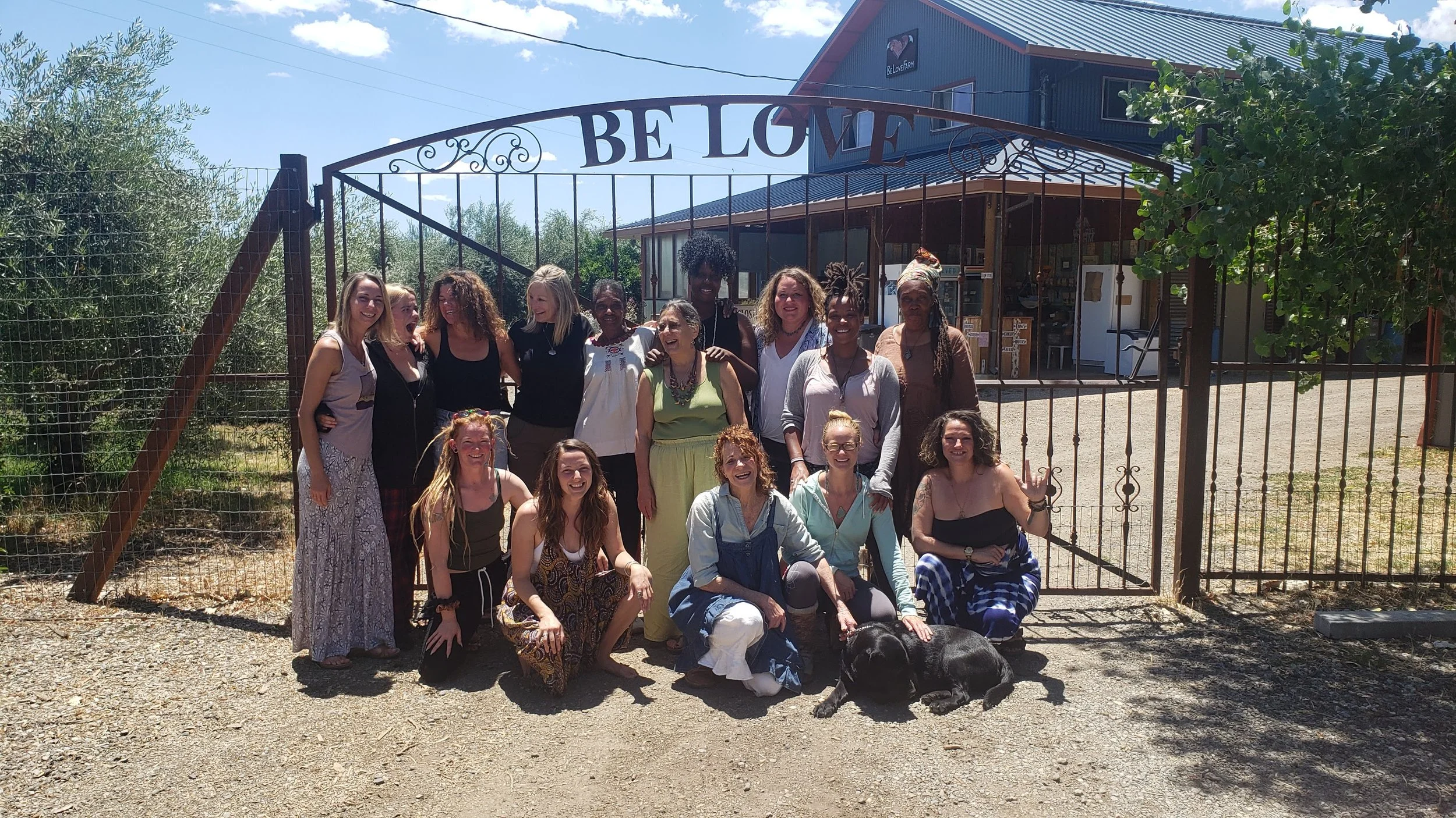 Group of women posing in front of a farm gate with a sign that says 'Be Love', some women crouching, some standing, with a black dog laying on the ground nearby, sunny day with blue sky and some clouds.