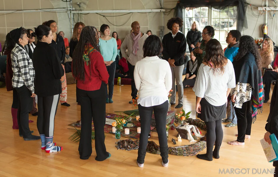 Diverse group of women gathered in a circle inside a tent, participating in a ceremony with flowers, candles, and offerings on the floor, while others sit and stand around observing. Taken at a WisdomWomen Visionary Gathering at Esalen Institute