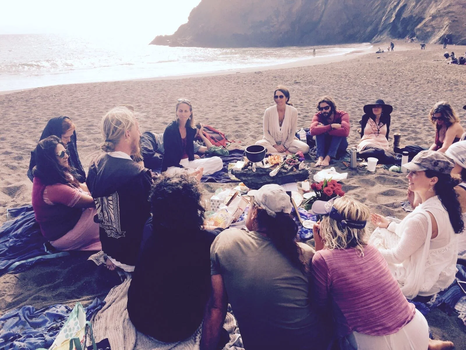 Group of people sitting in a circle on a beach, having a picnic near the water, with cliffs in the background.